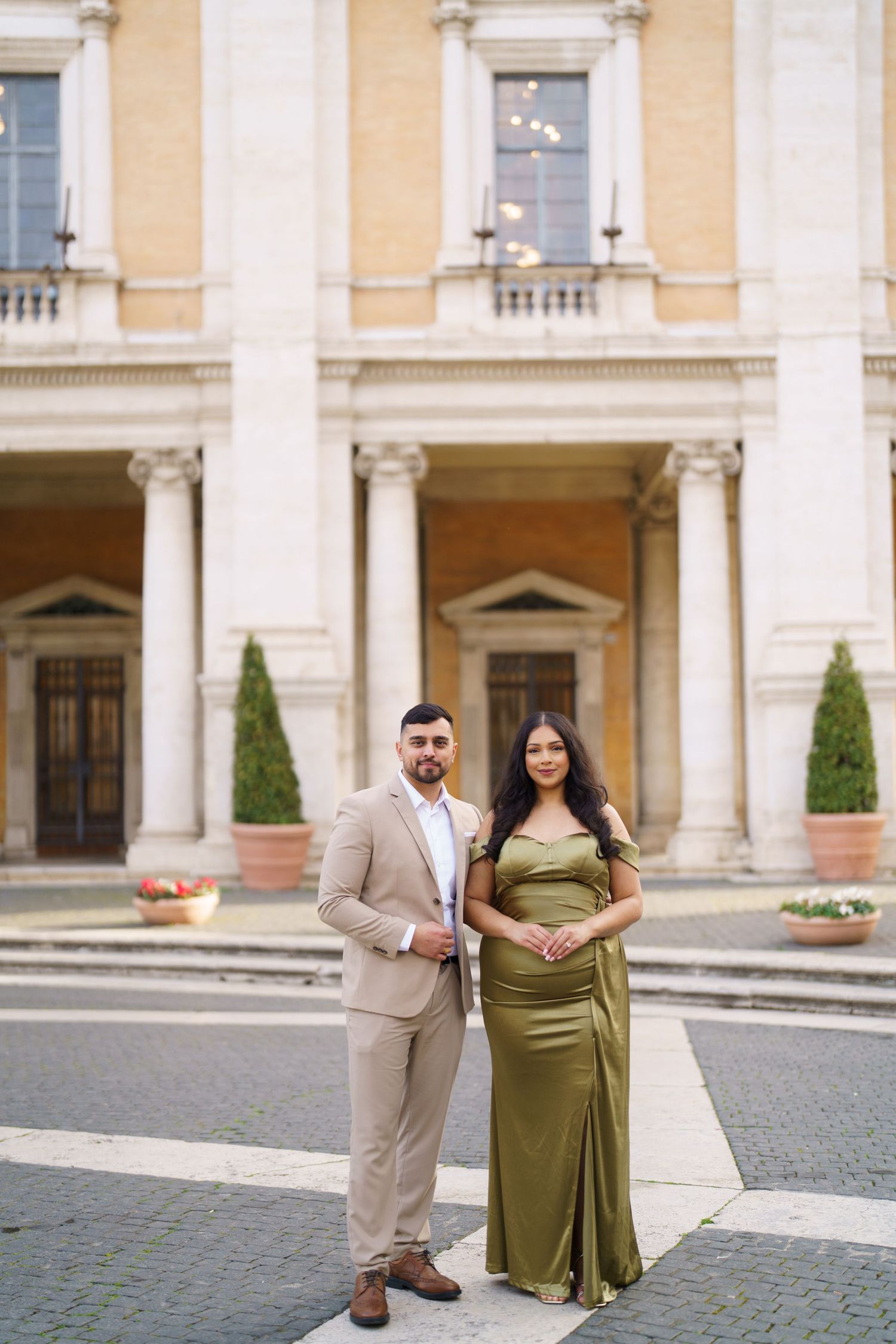 Elegant couple posing in front of a classical building, man in beige suit, woman in olive green gown.