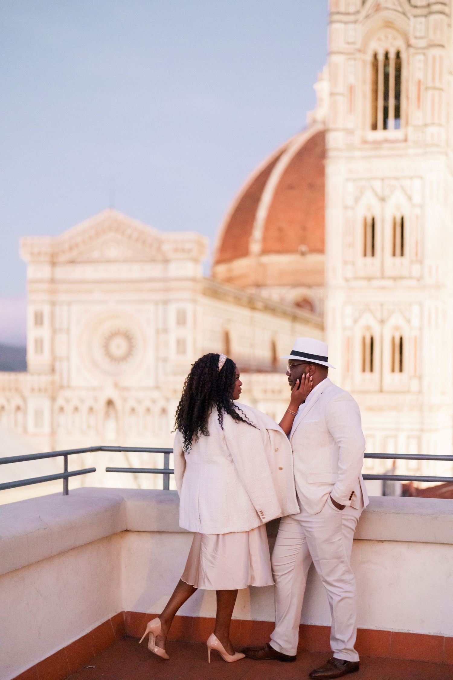 Couple in white outfits on a rooftop terrace with Florence Cathedral dome backdrop at dusk.
