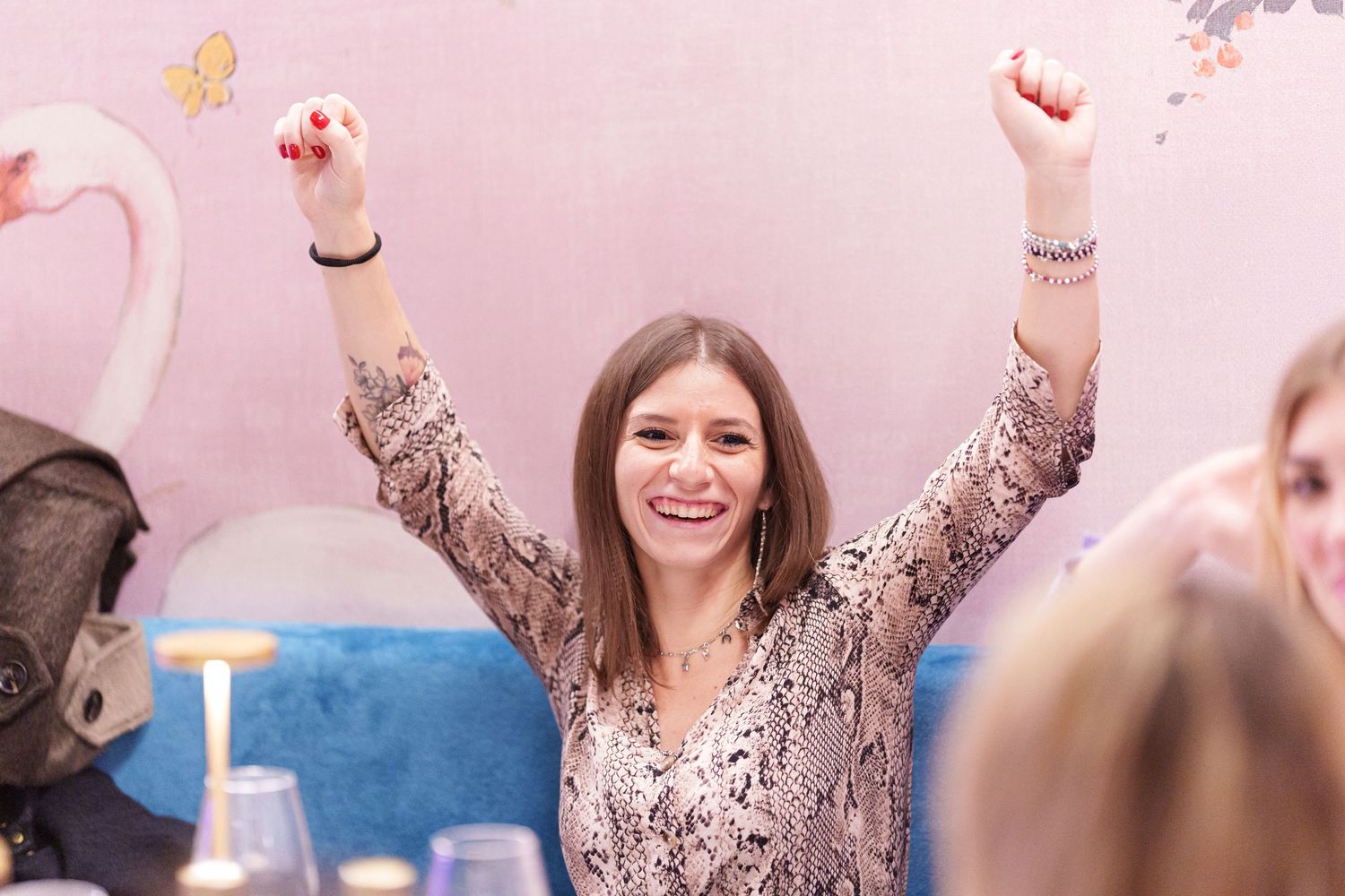 Excited young woman raising both arms in victory, smiling joyfully at a pink-themed indoor gathering.