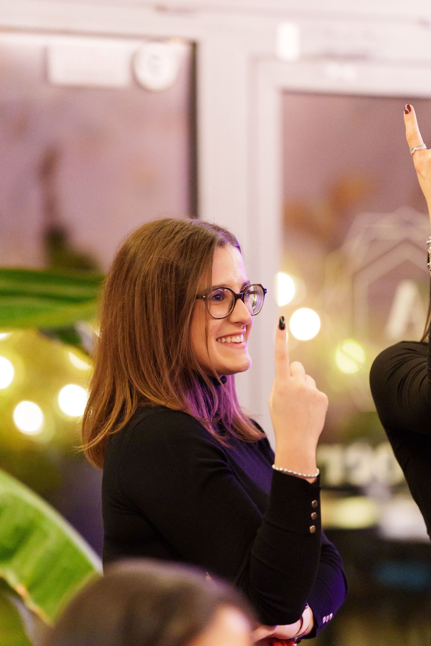 Smiling woman with glasses and purple hair highlights raises finger at a festive indoor event with balloons.