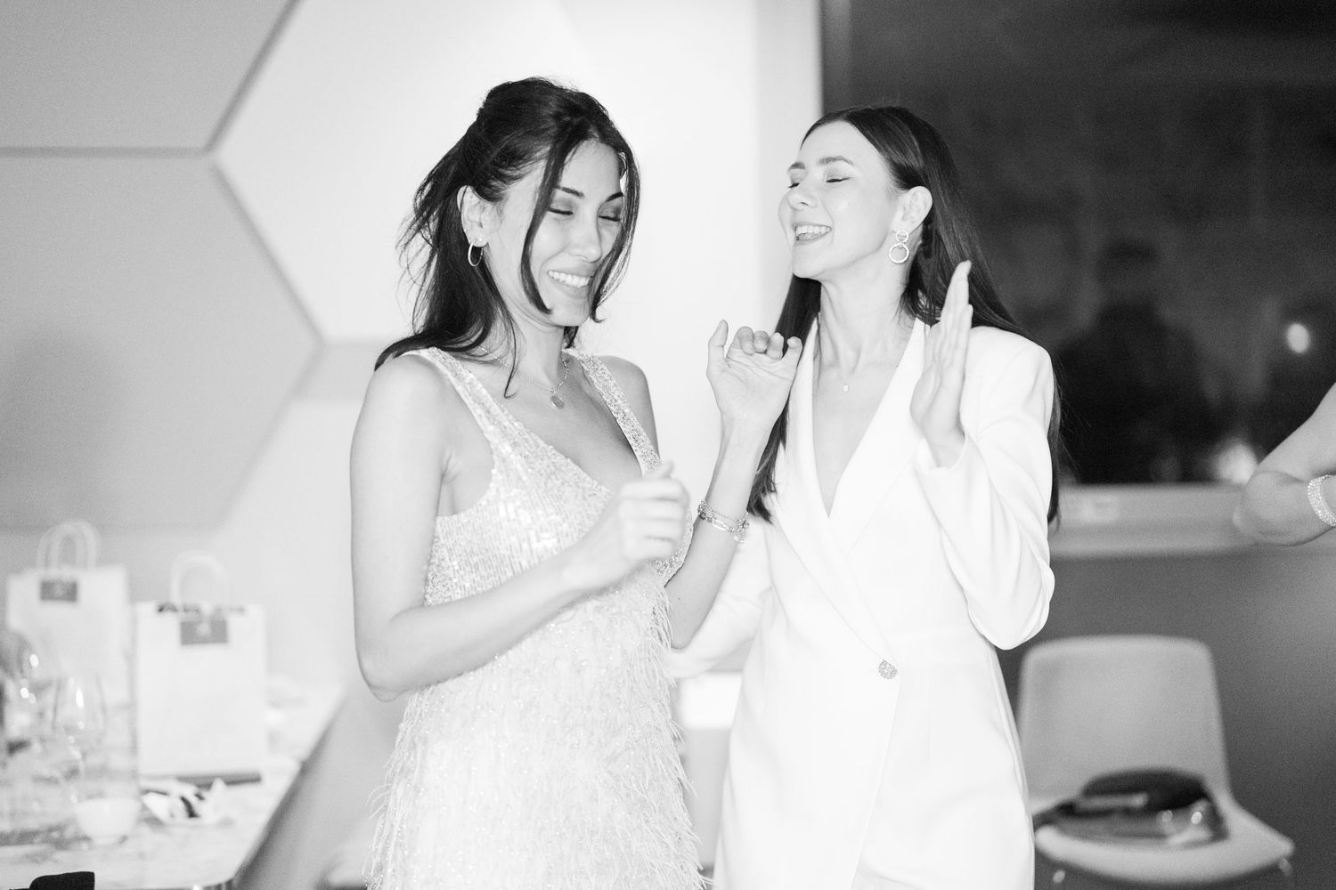 Two elegant women in white outfits laughing together at a stylish event in black and white photo.