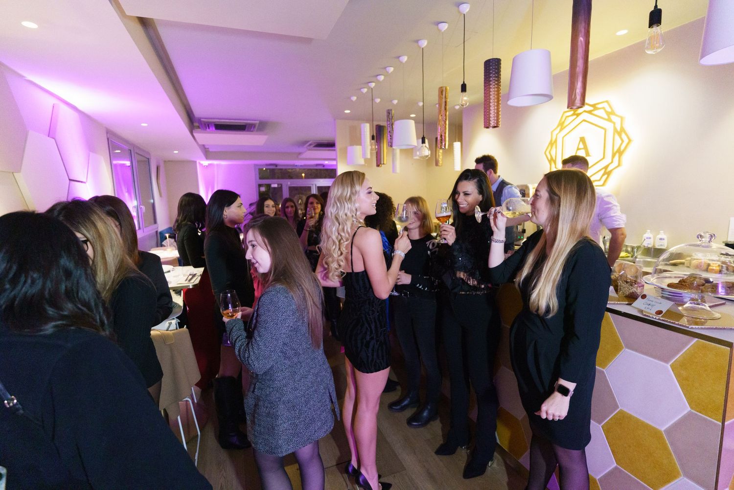 Elegant indoor cocktail party with guests mingling, wearing black attire, under warm pendant lights in a modern venue.