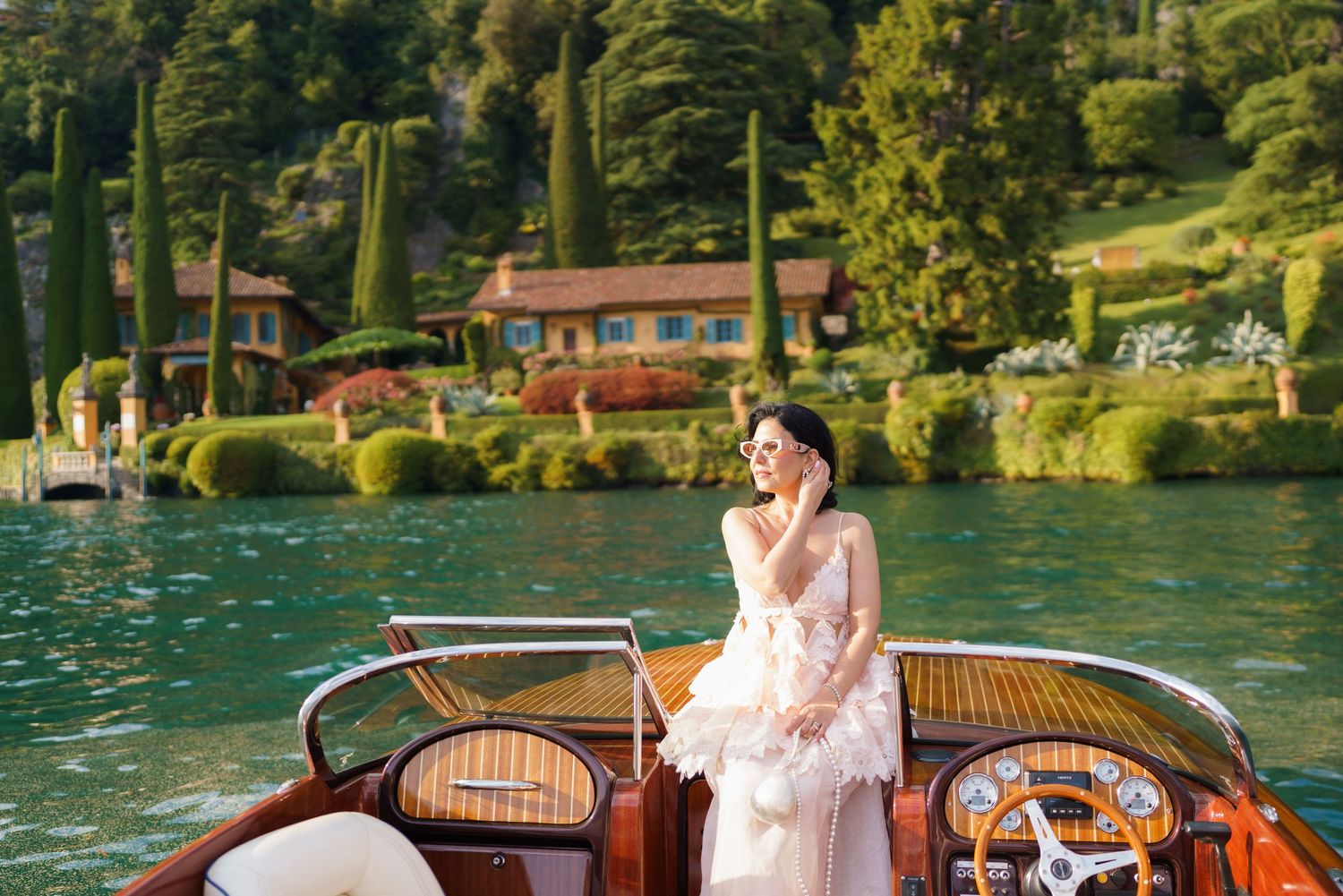Bride in a pink feathered gown sits on a wooden boat on Lake Como, Italy, with lush green hills and a villa behind her.
