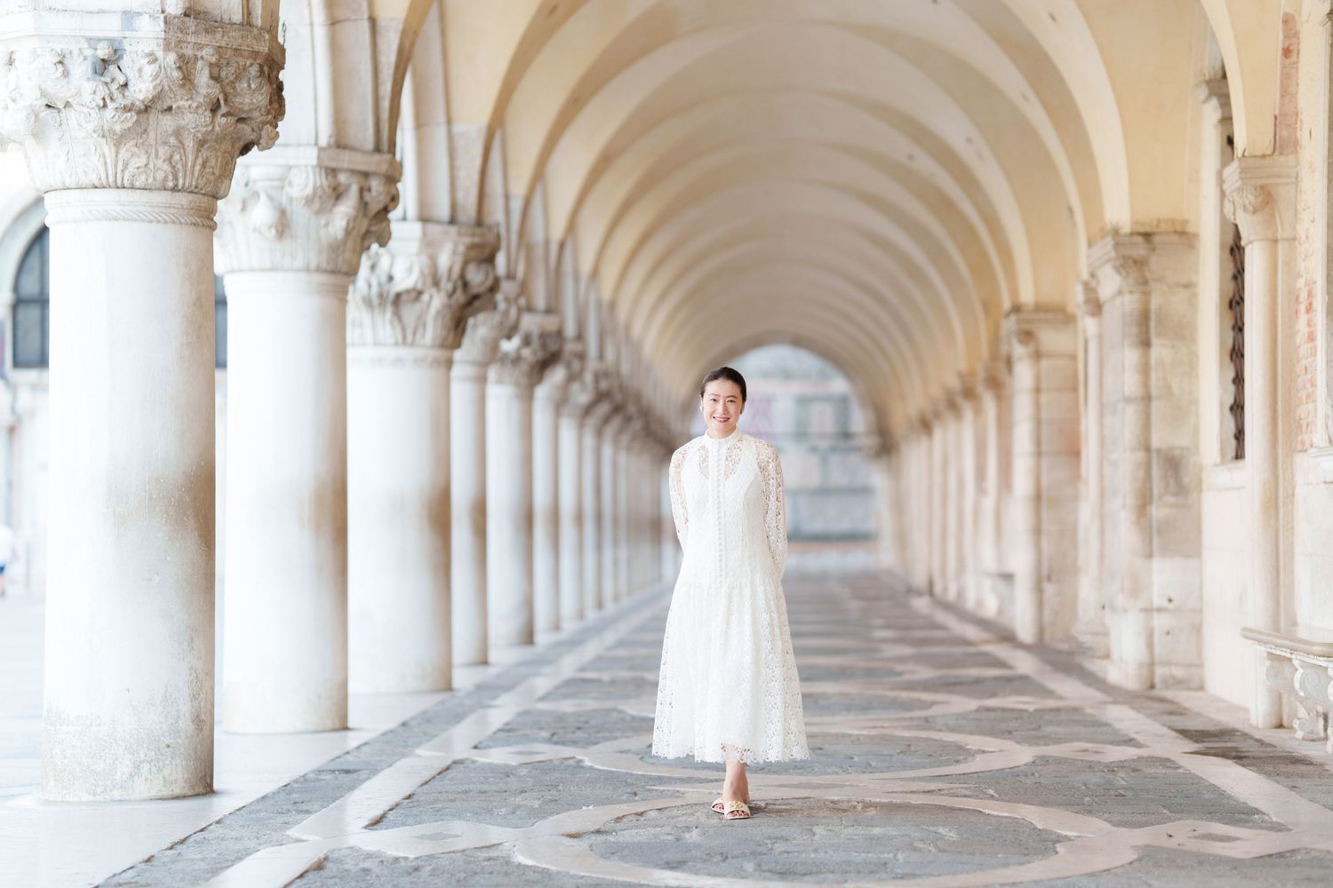 Woman in white midi dress walking under elegant arched colonnade in Venice, Italy.