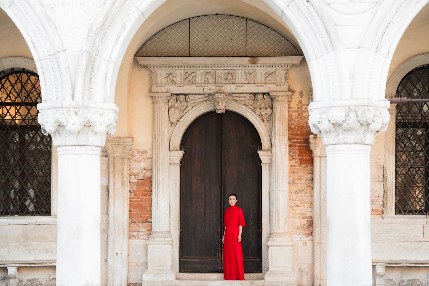 Woman in red dress standing under white marble arches before ornate wooden door in Venice, Italy.