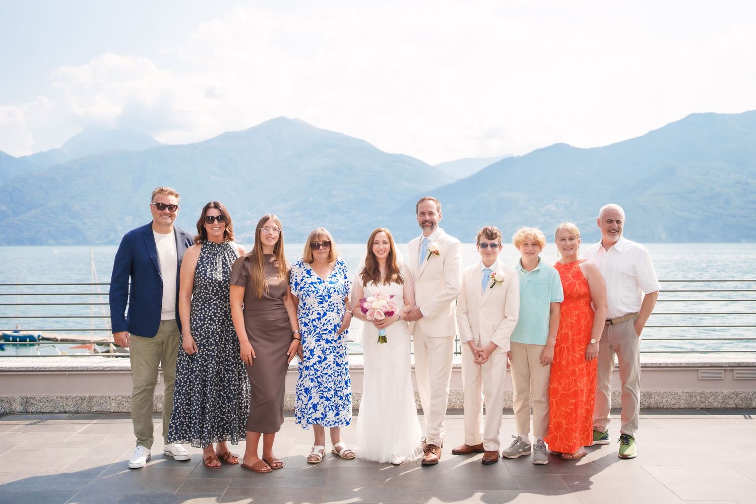 Wedding party poses on lakeside terrace with mountain backdrop, bride in white gown holding bouquet with family como lake