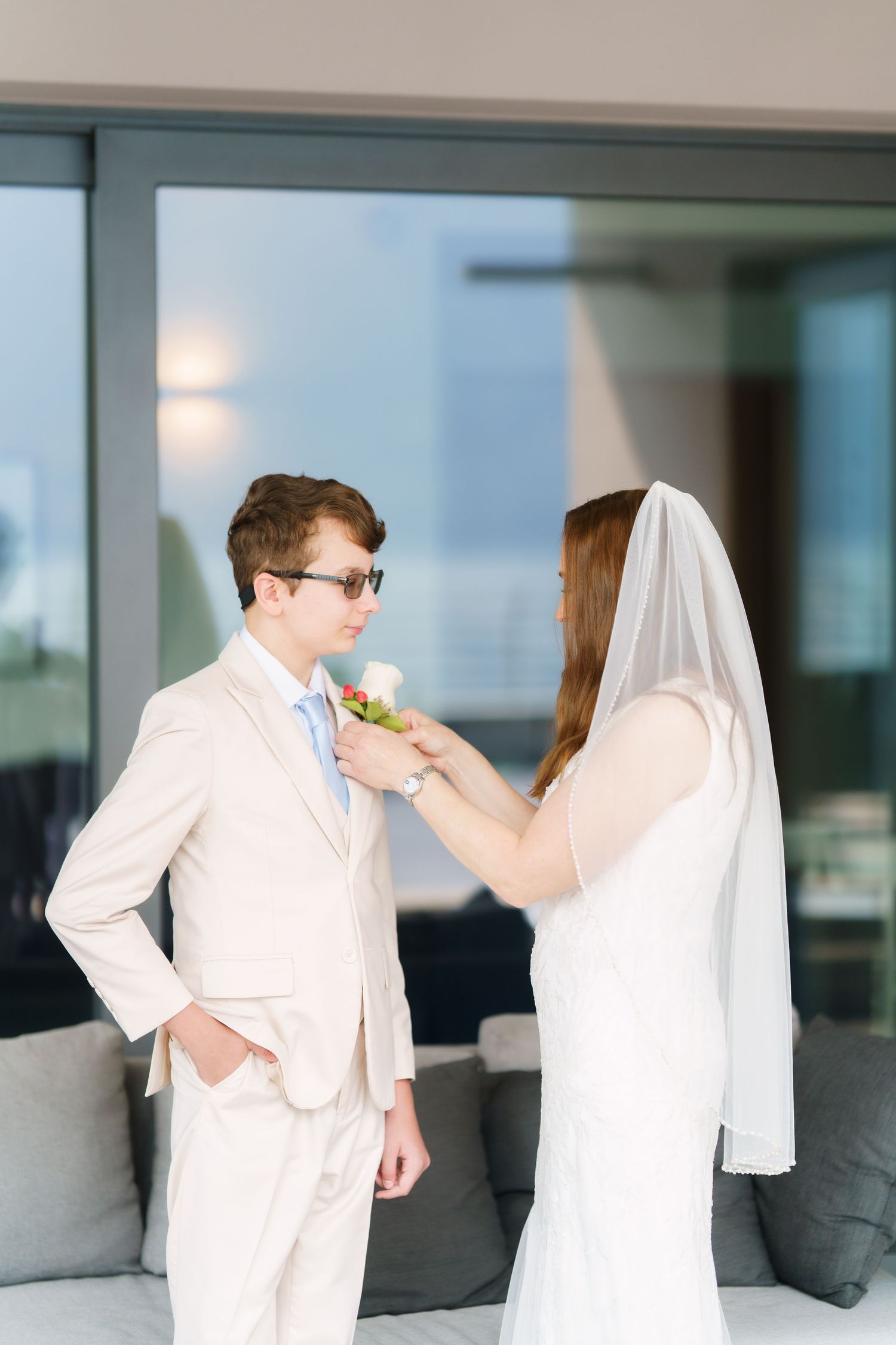 Bride in white gown and veil pins boutonniere on groom in beige suit with sunglasses at modern venue.