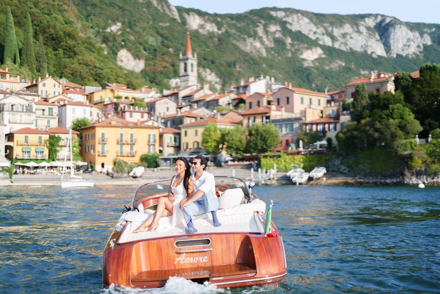 Two women on a wooden boat cruising Lake Como with colorful Italian village buildings and green hills in background.