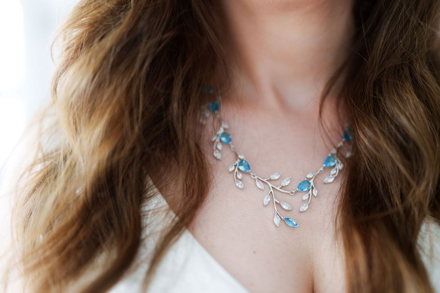 Woman wearing an elegant silver branch necklace with blue gemstones and white floral accents on a white outfit.