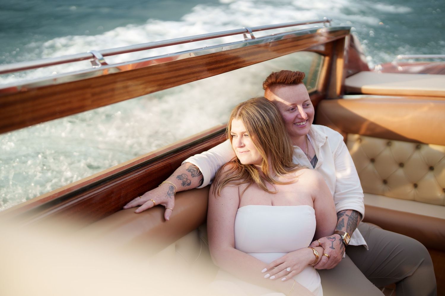 Couple relaxing on a luxury wooden boat, man with tattoos embracing woman in white top on tufted leather seat.
