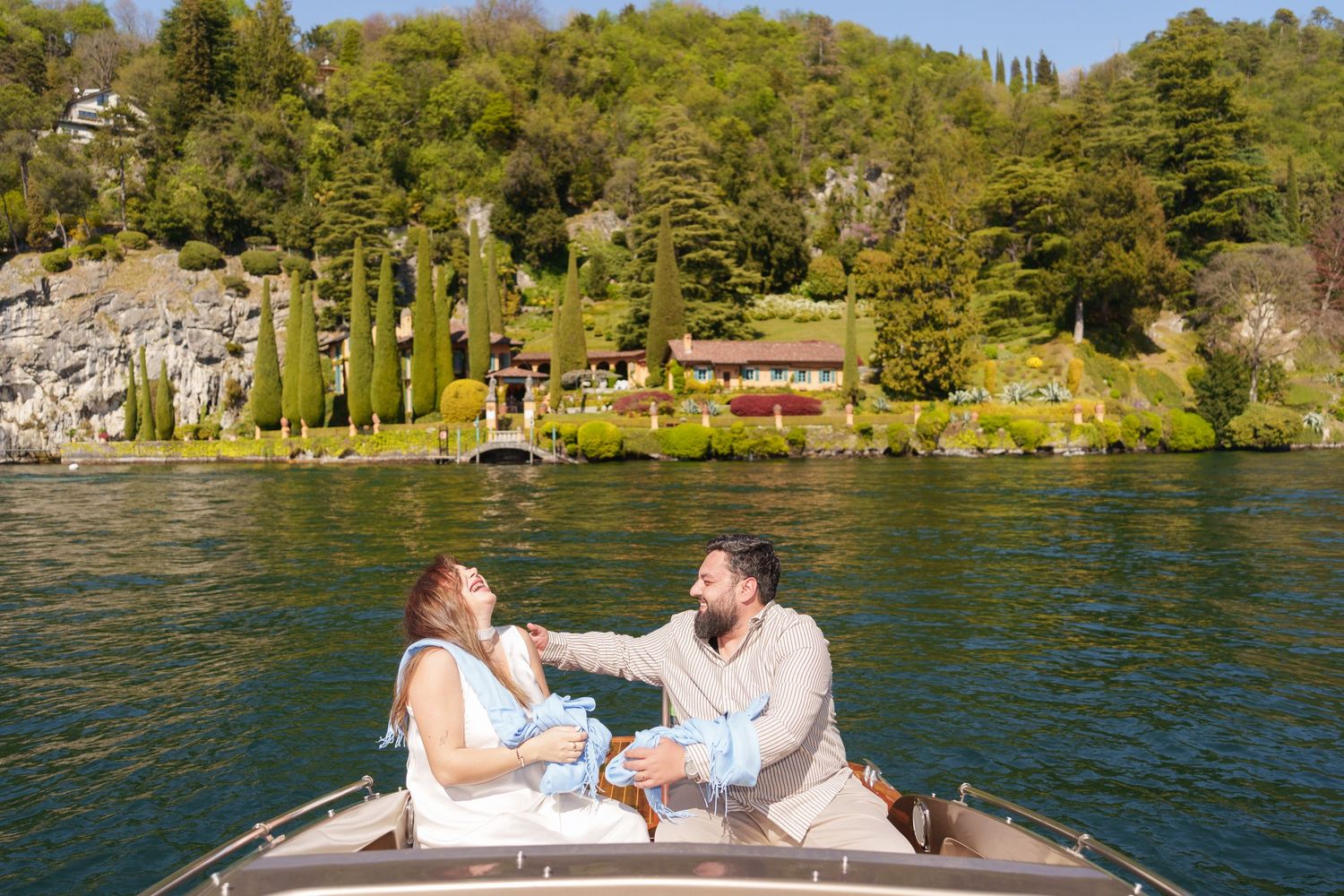 Couple sitting together on a boat enjoying a scenic lake view with lush green hillside and red villa in background.