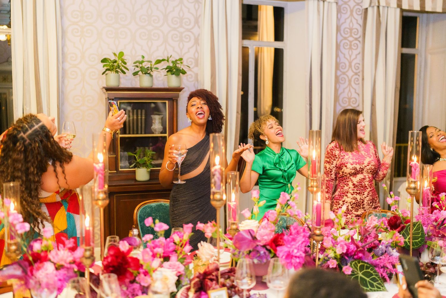 Women laughing and celebrating at a colorful floral dinner party with candles and pink flowers.
