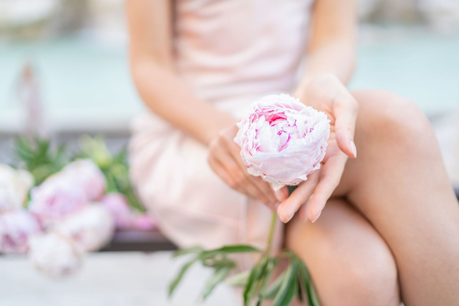 Woman in pink dress holding a delicate white and pink peony flower with soft blurred floral background.