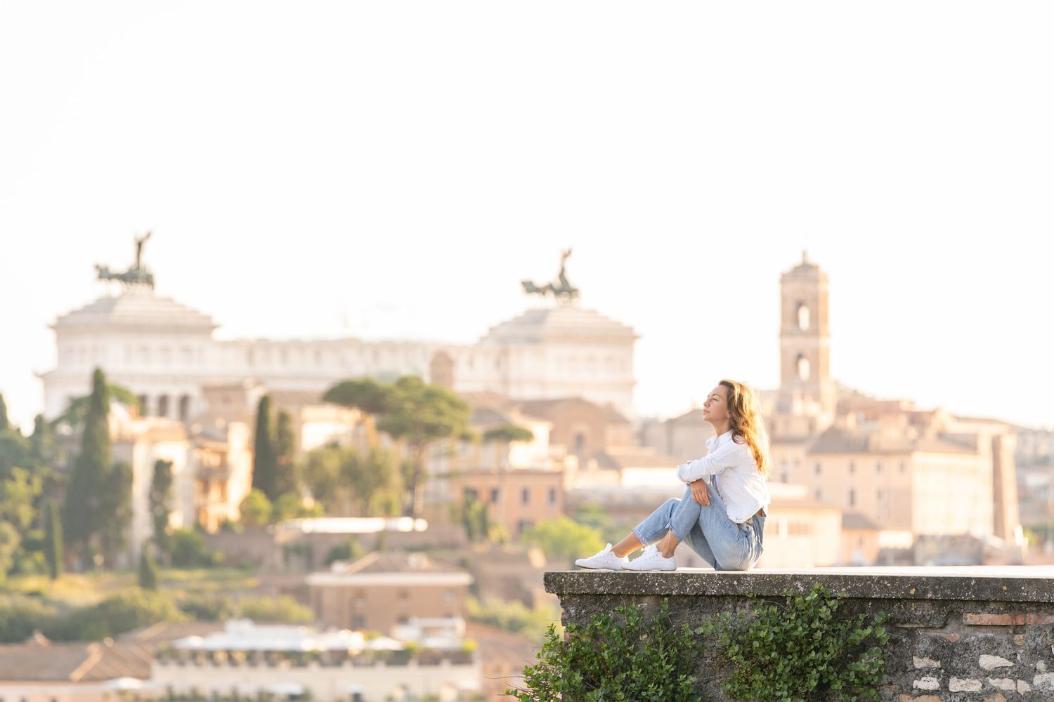 Woman sitting on ancient stone wall overlooking Rome skyline with historic monuments and church towers in soft golden light.