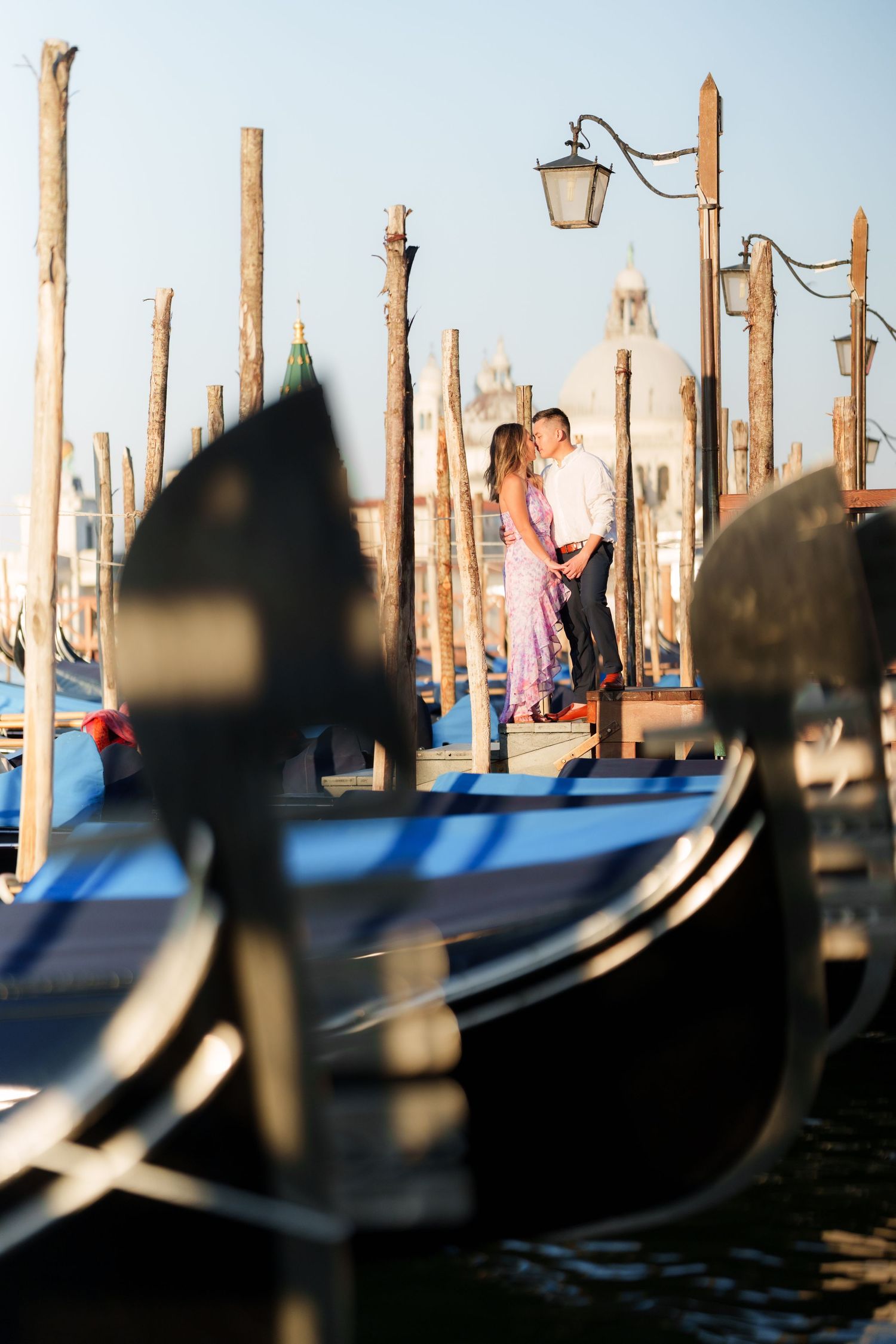 Romantic couple embracing among gondolas and wooden poles at Venice waterfront with church in background.