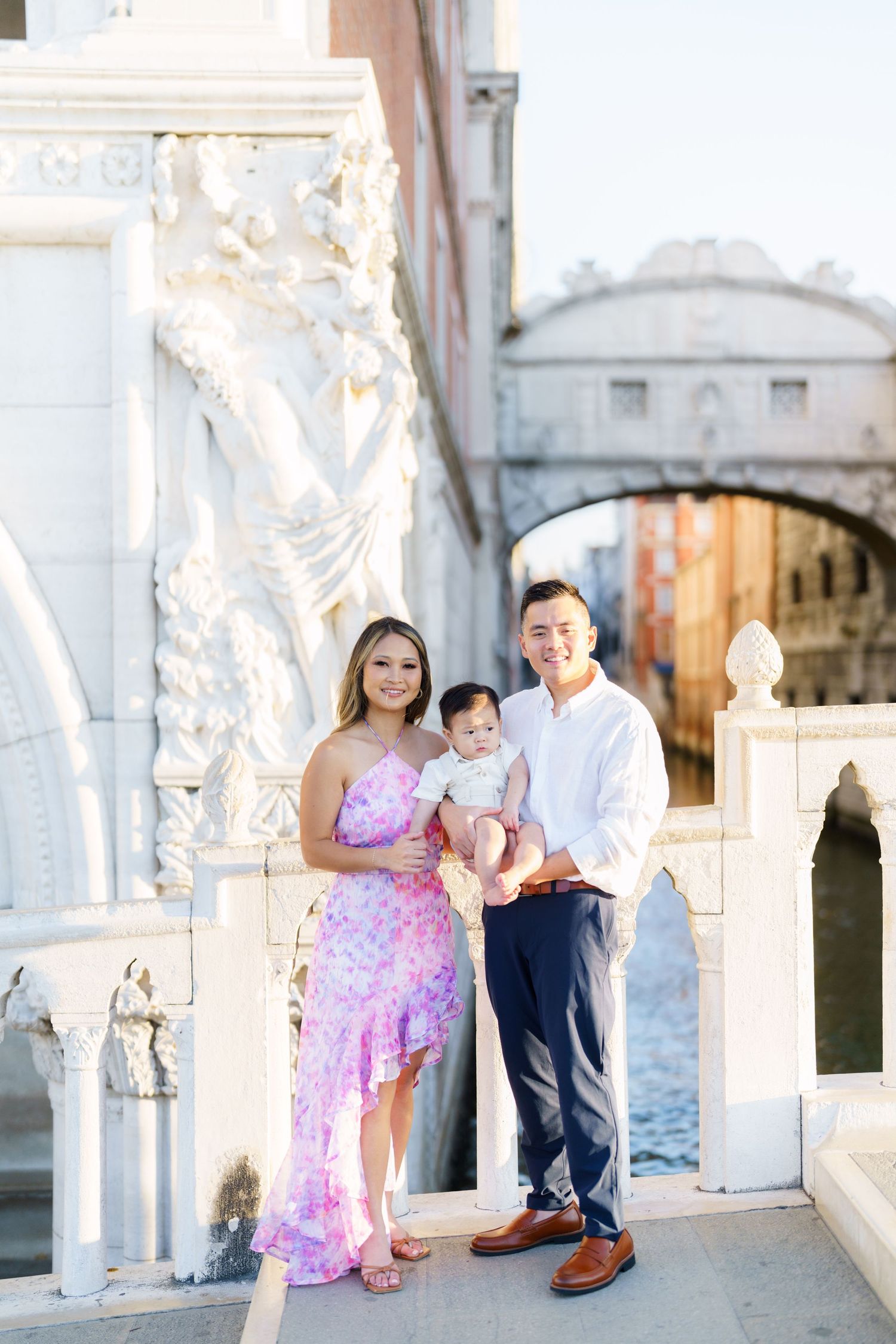 Family of three posing near the Bridge of Sighs in Venice, Italy, with a canal and historic architecture in the background.