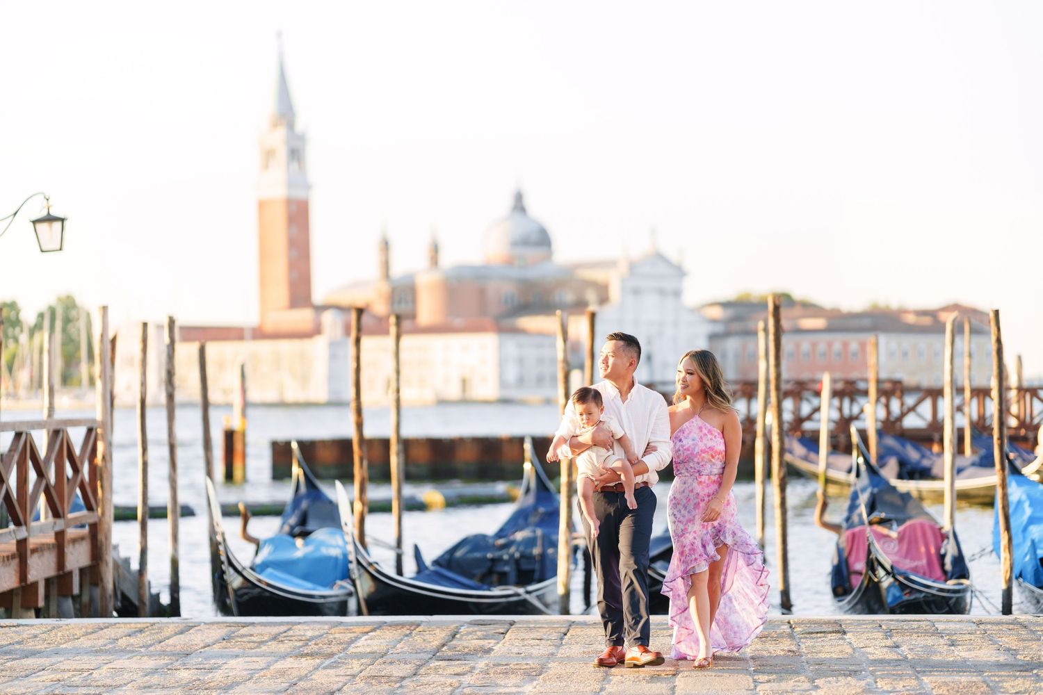 Family with baby posing by gondolas on Venice waterfront with San Giorgio Maggiore church in background.