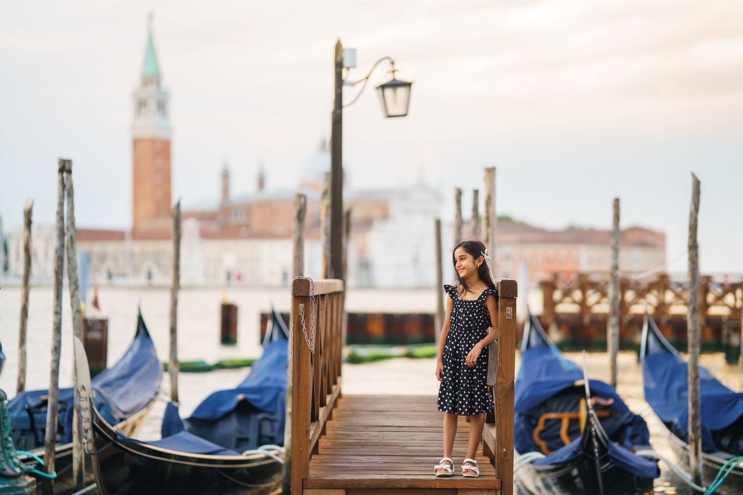 A woman in a polka dot dress stands on a wooden dock with gondolas in Venice, Italy.