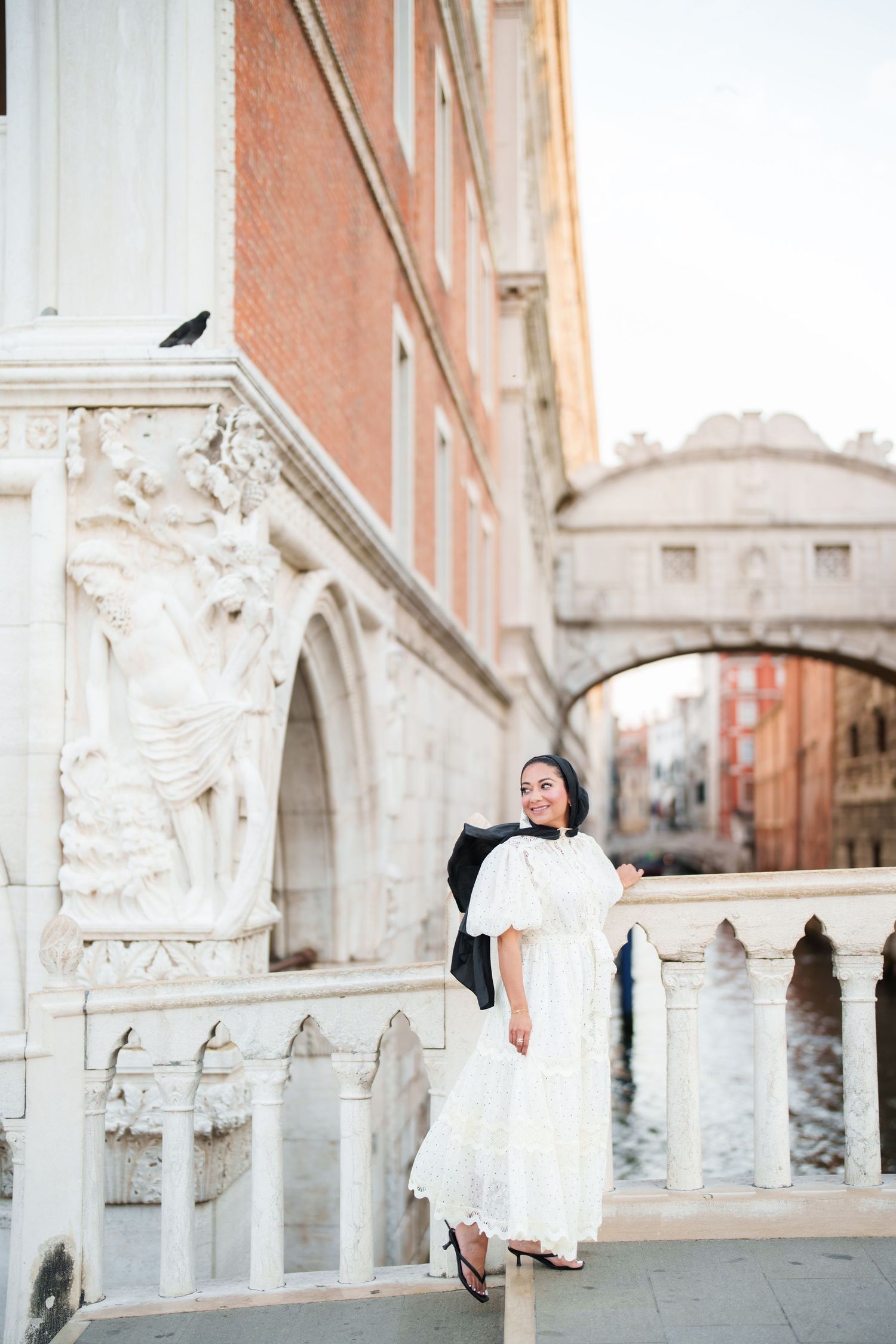 Woman in white dress poses near ornate marble columns with Venice's Bridge of Sighs and canal in background.