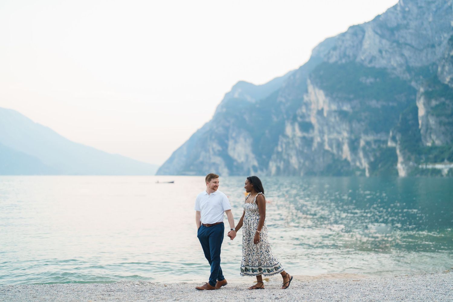Interracial couple strolling hand in hand along Lake Garda shore with alpine mountains.