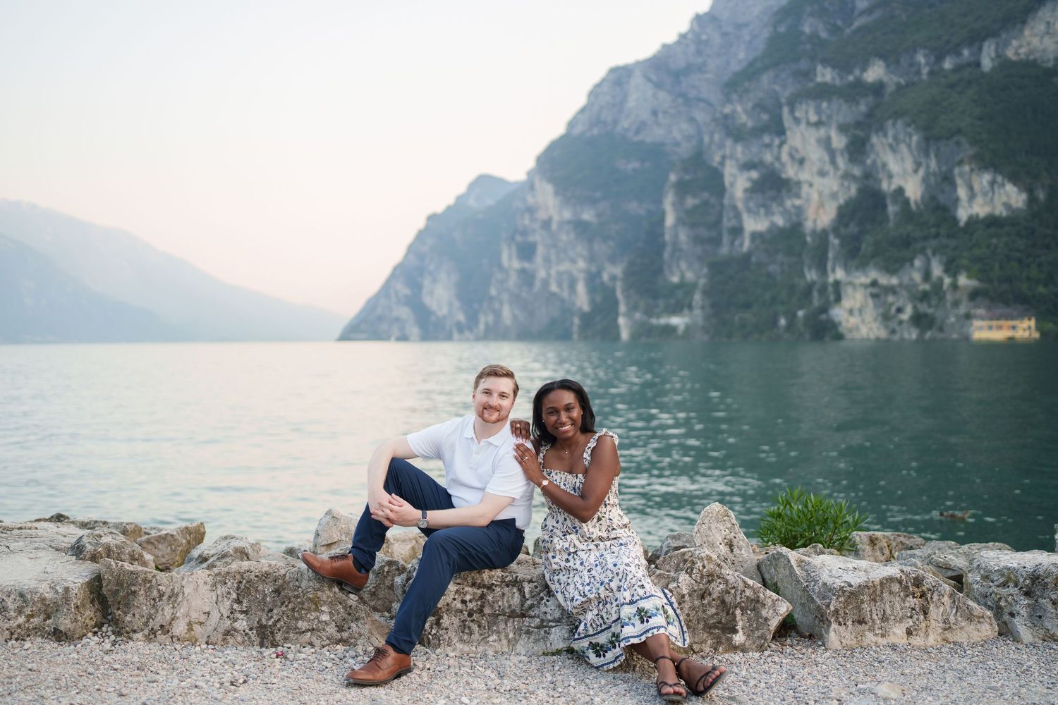 Smiling interracial couple sitting together on rocks with turquoise Lake Garda and cliffs in the background.