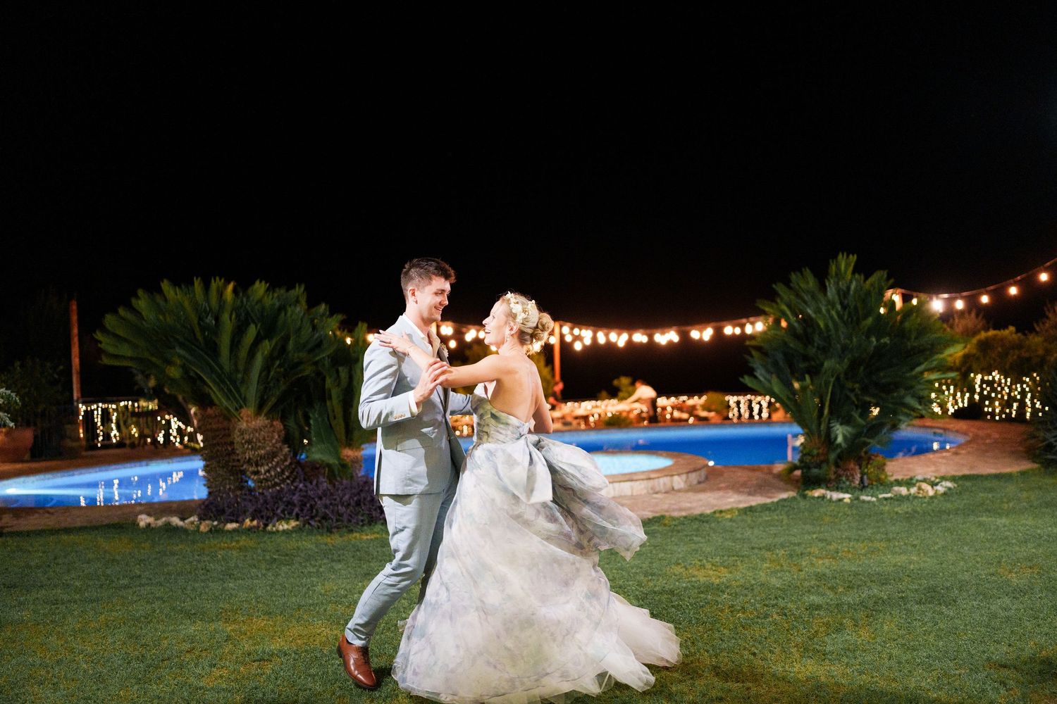 Bride and groom share first dance outdoors at night near illuminated pool with string lights at elegant wedding reception.