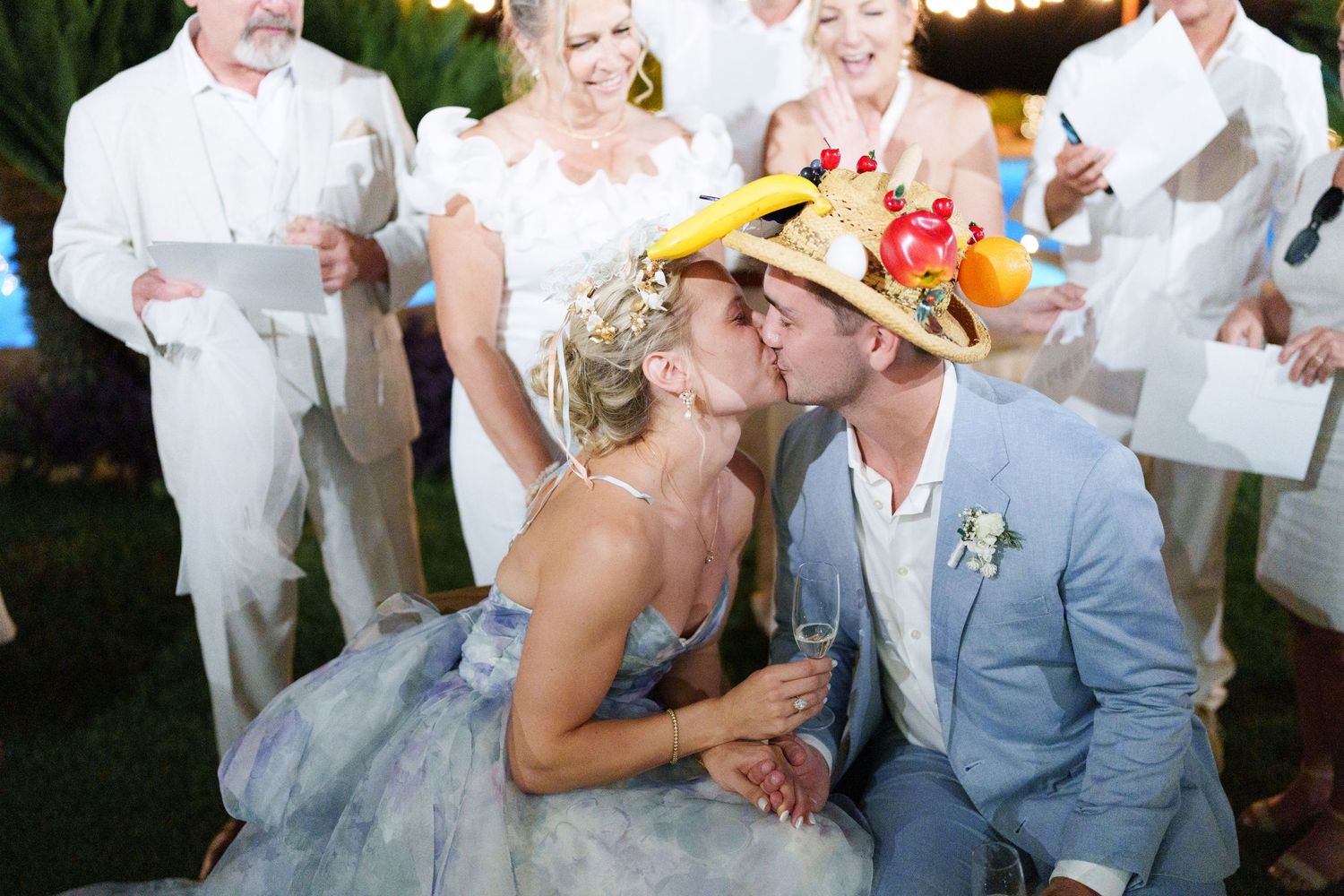 Bride and groom sharing a kiss wearing colorful fruit hats at an outdoor wedding reception with guests in background.