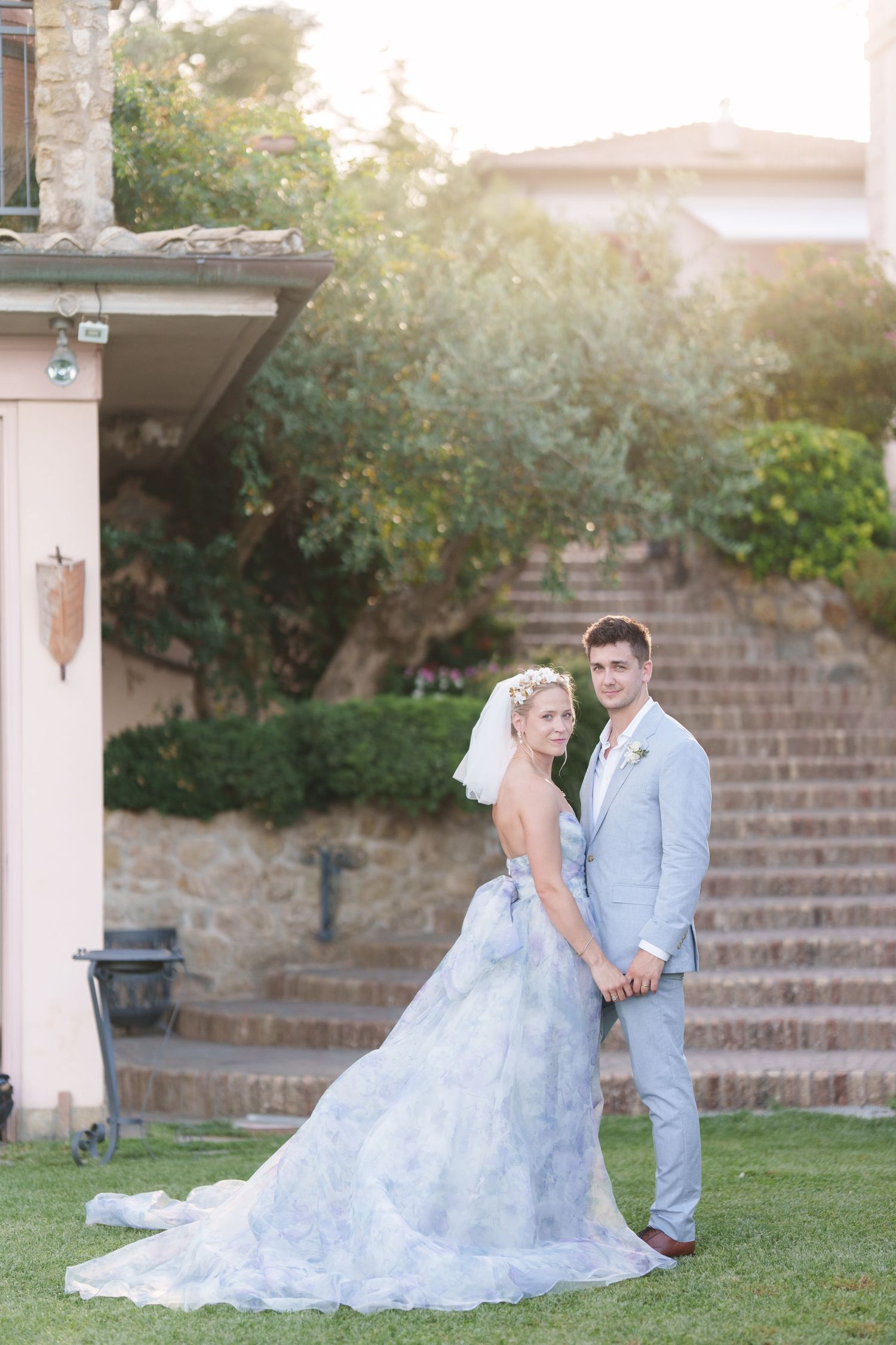 Bride in blue lace gown and groom in light gray suit pose together on green lawn at golden hour.