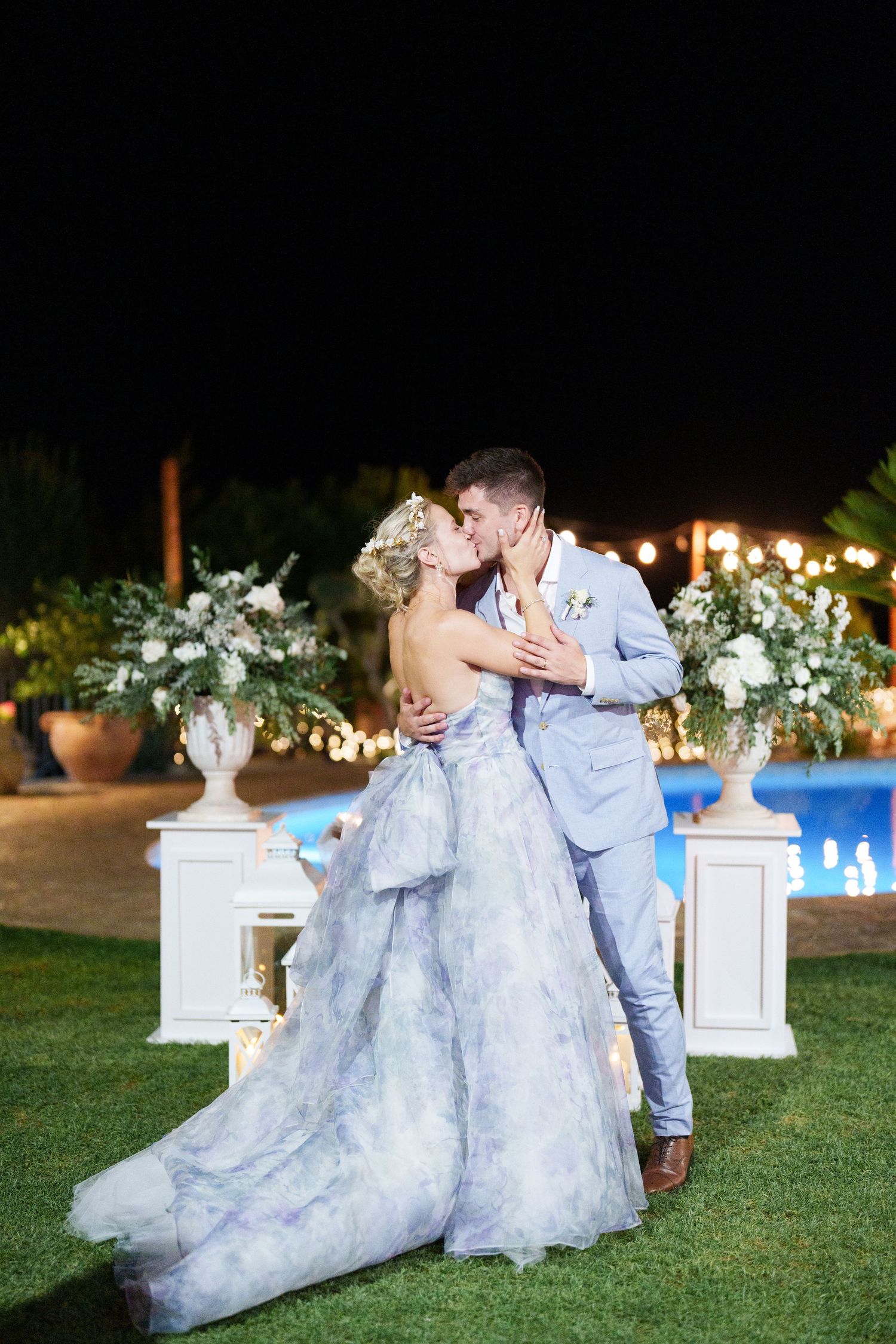 Bride in blue gown and groom in light suit share a romantic kiss by a pool with floral arrangements at night.