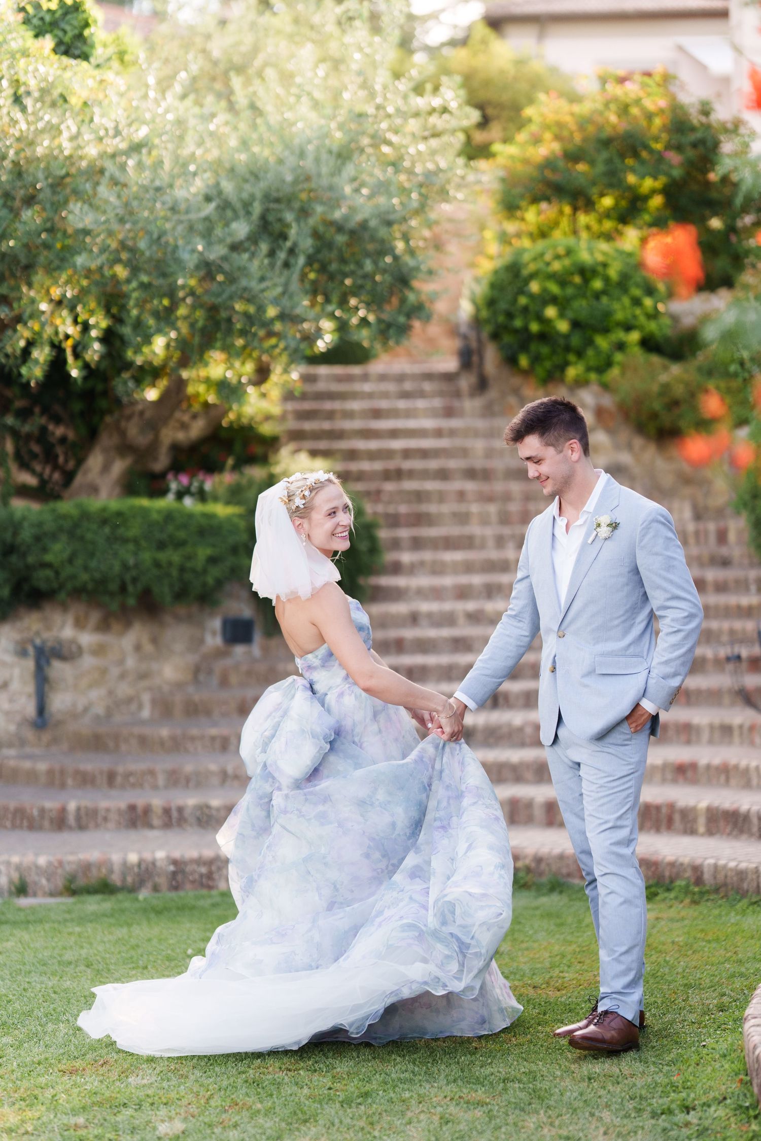 A bride in a blue floral gown holds hands with her groom in a light gray suit on a green lawn with stone steps behind them.