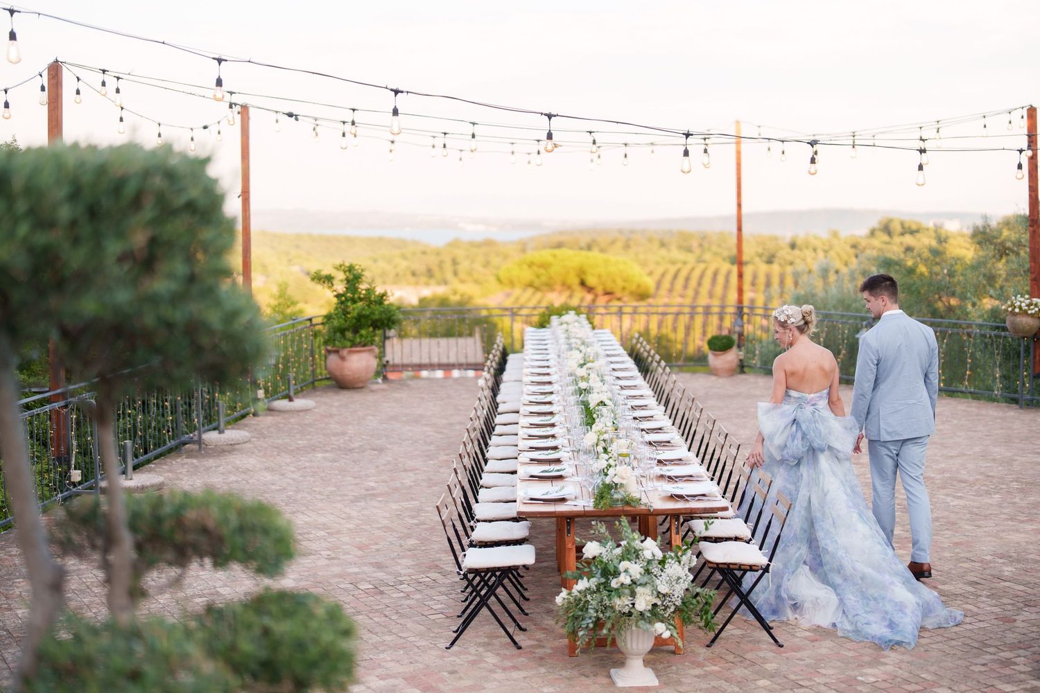 Couple walks toward long outdoor reception table with string lights at a vineyard wedding venue at sunset.