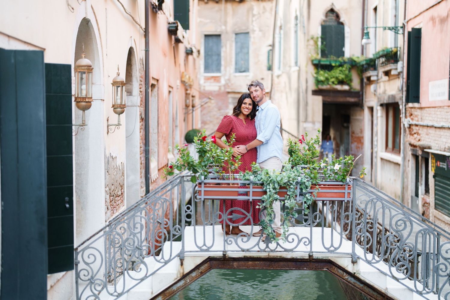 Couple embracing on ornate bridge over Venice canal surrounded by colorful flowers and historic buildings.