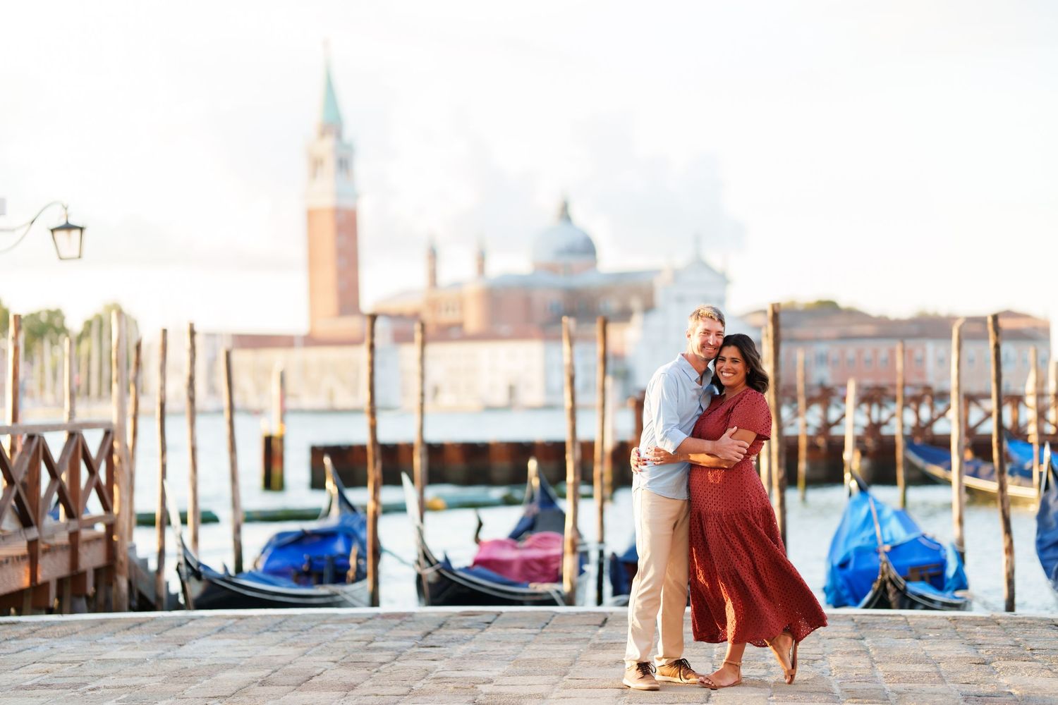 Couple embracing romantically by Venice gondolas with San Giorgio Maggiore church in background at sunrise.
