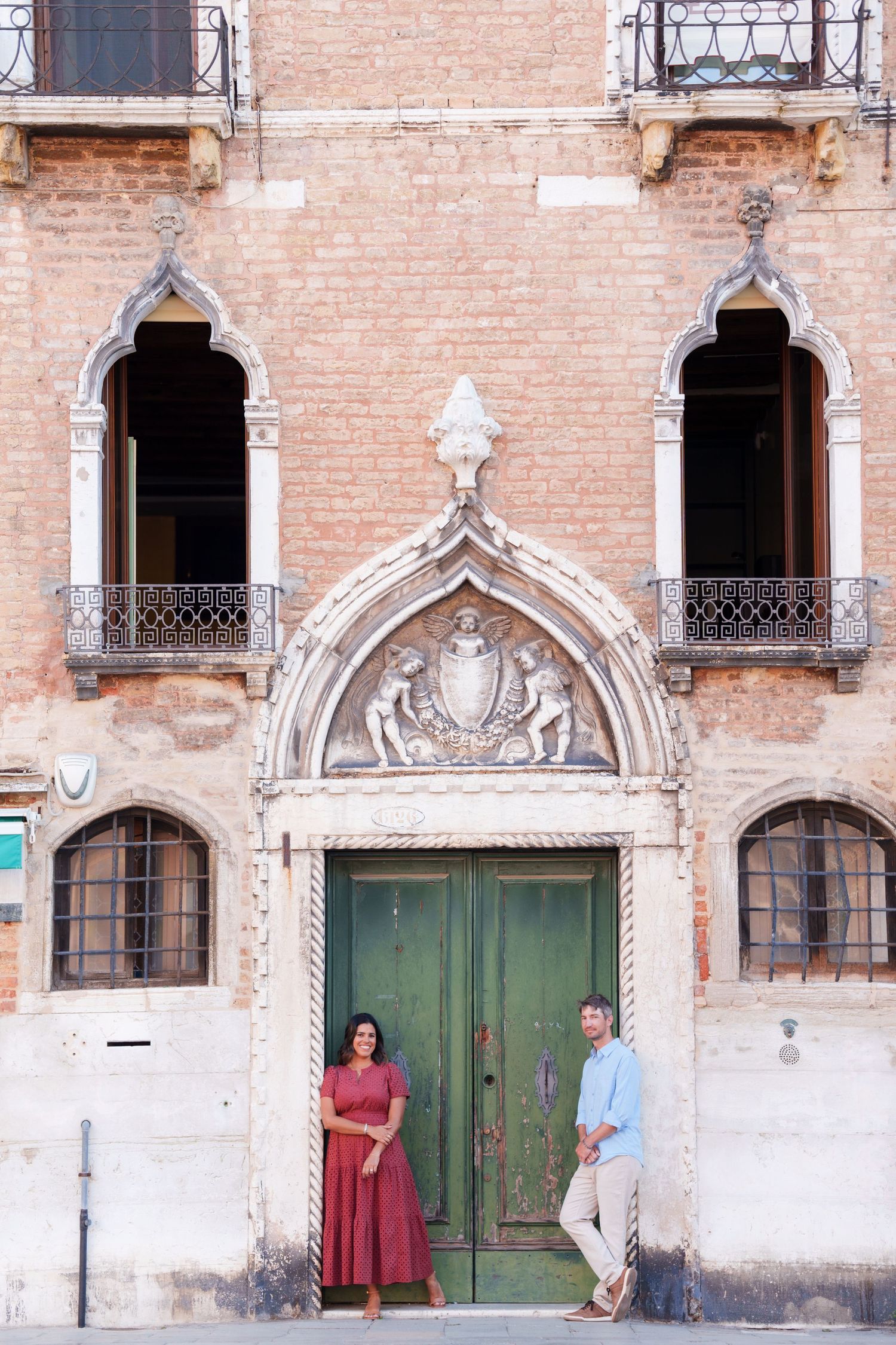 Couple posing by ornate Gothic green door of historic Venetian building with decorative arch and iron balconies.
