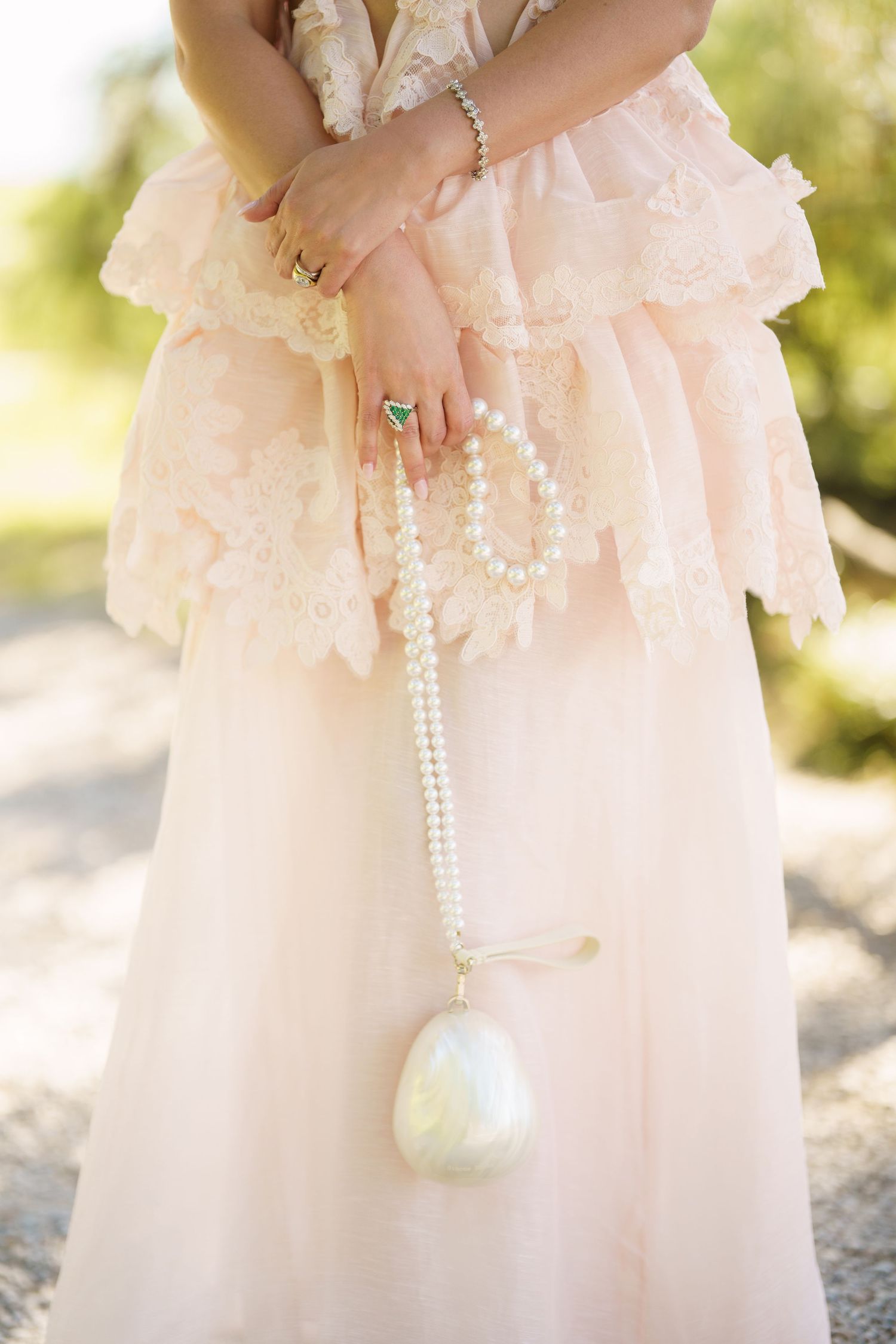 Woman in blush lace dress holding pearl necklace with large baroque pearl pendant, wearing emerald ring.