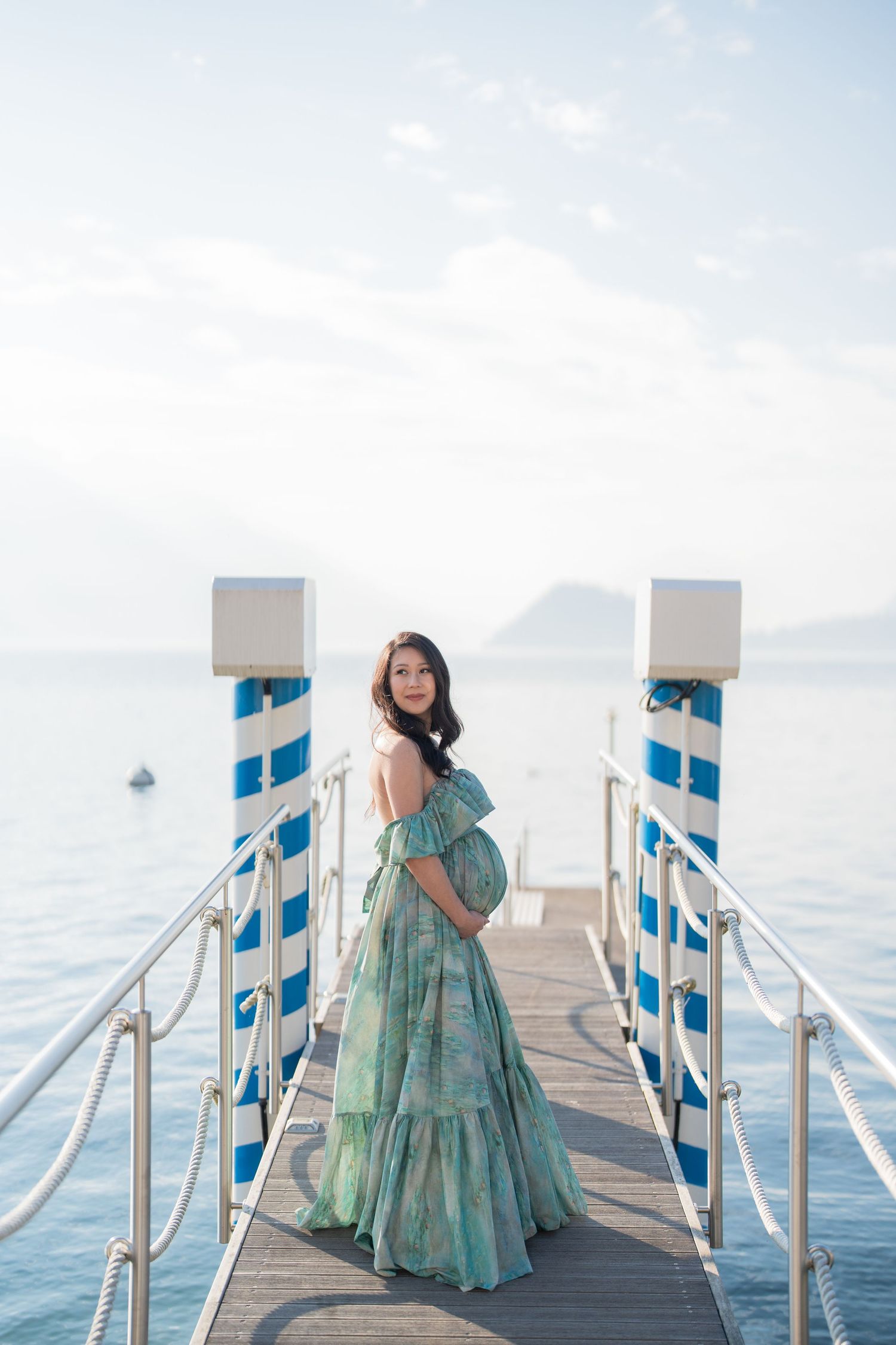 Pregnant woman in green floral off-shoulder gown standing on a pier overlooking calm blue ocean waters.