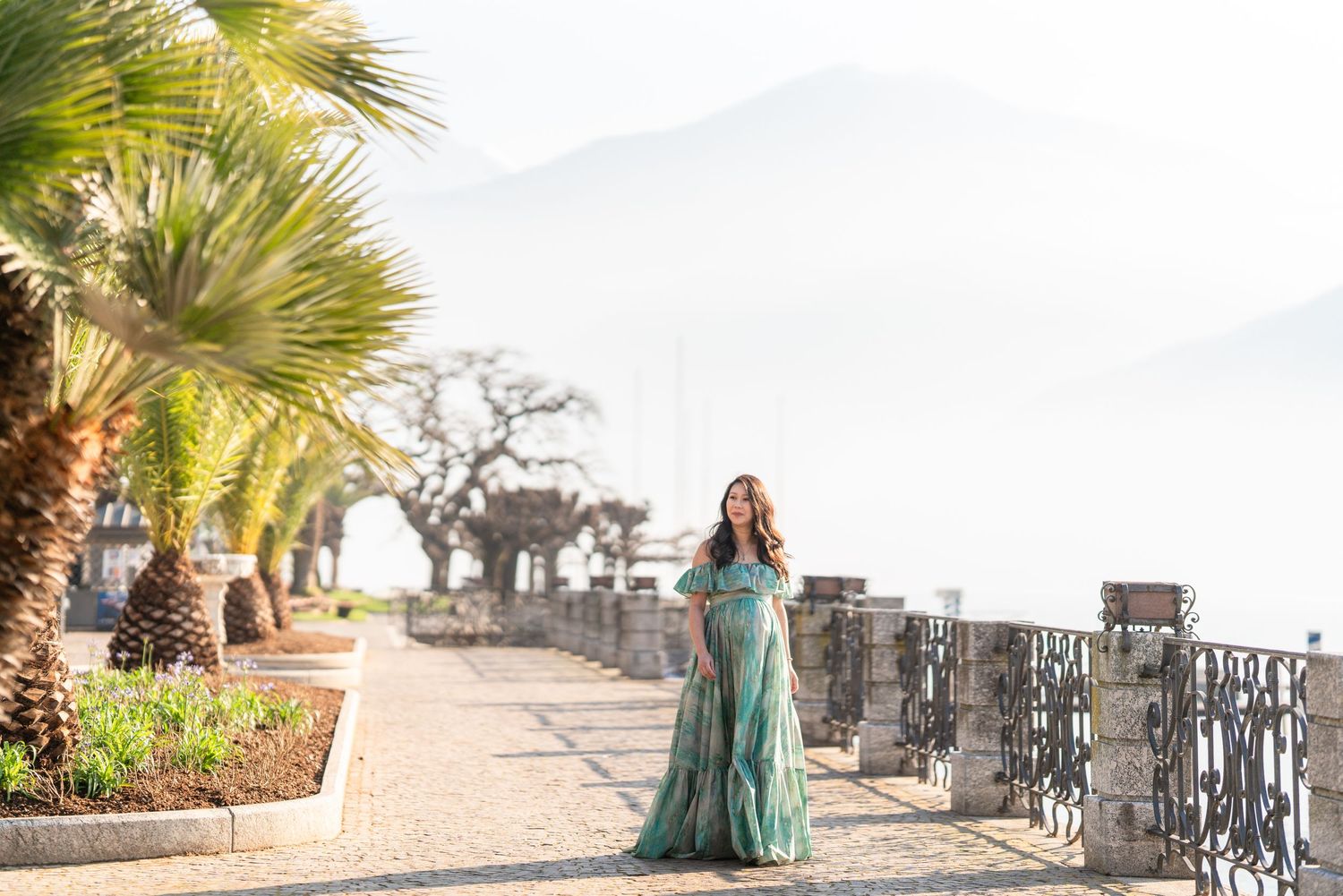 Woman in teal gown on scenic lakeside promenade with palm trees and misty mountains in background.
