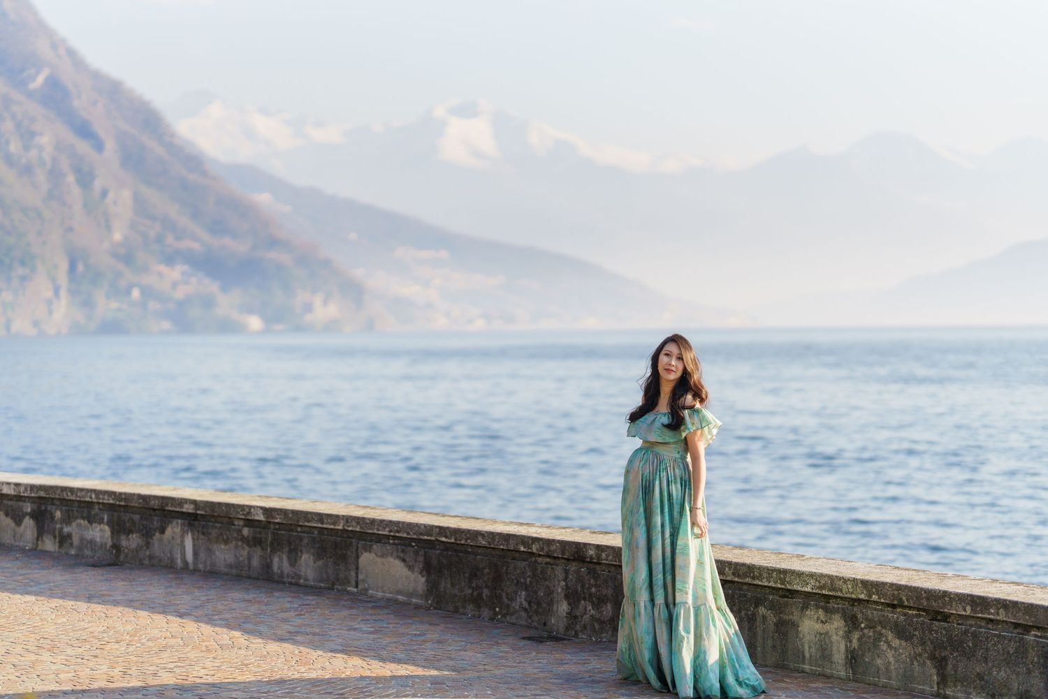 Pregnant woman in teal gown poses on lakeside promenade with misty mountains and serene blue water backdrop.