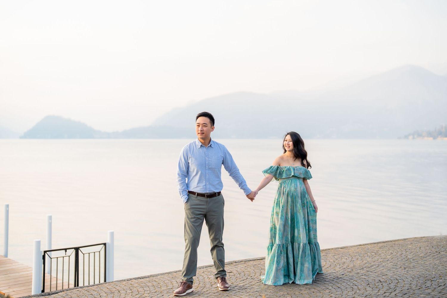 Couple holding hands on a lakeside promenade with misty mountains in the background, woman in a flowing teal dress.