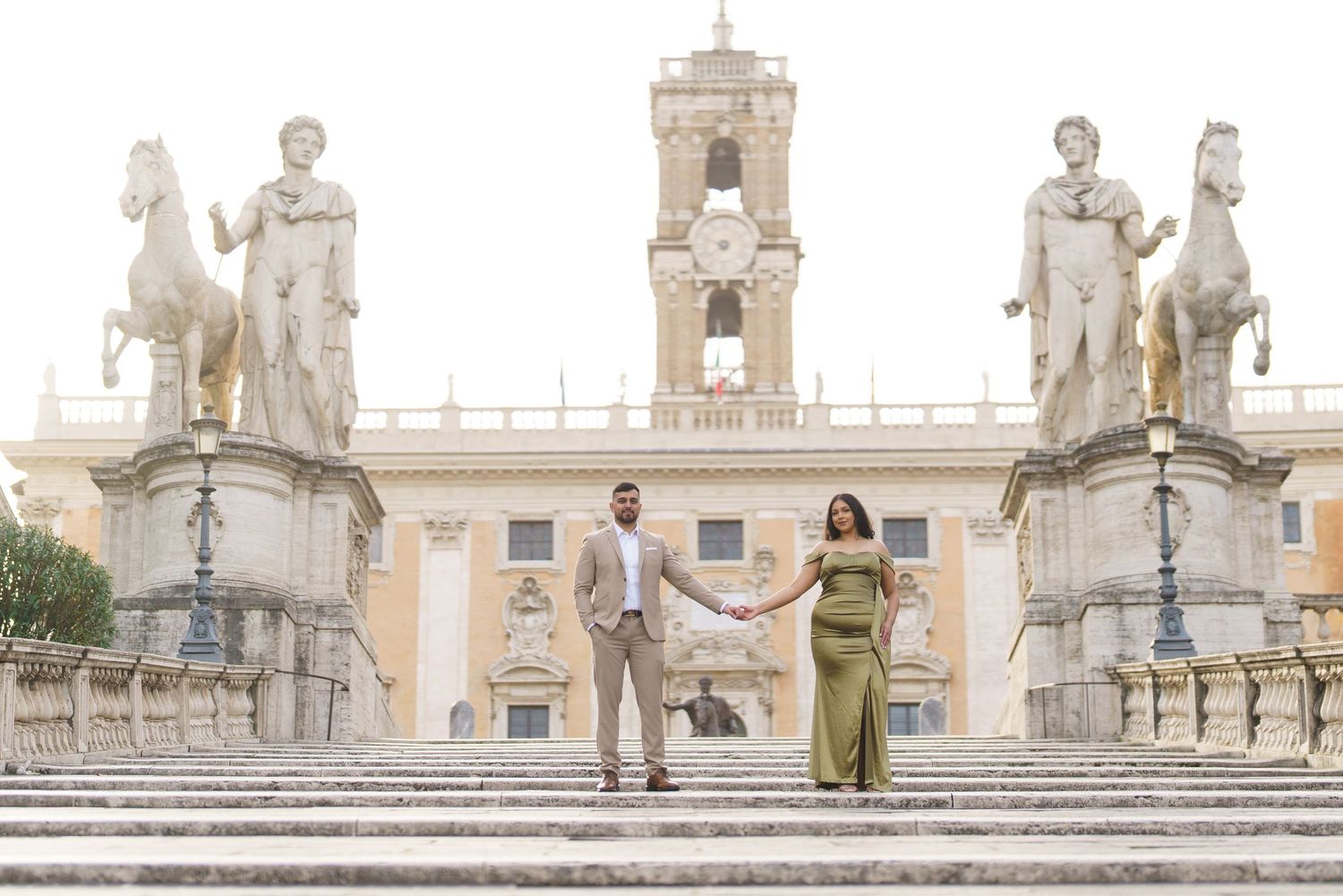 Couple holding hands on steps at Piazza del Campidoglio in Rome, flanked by classical statues.