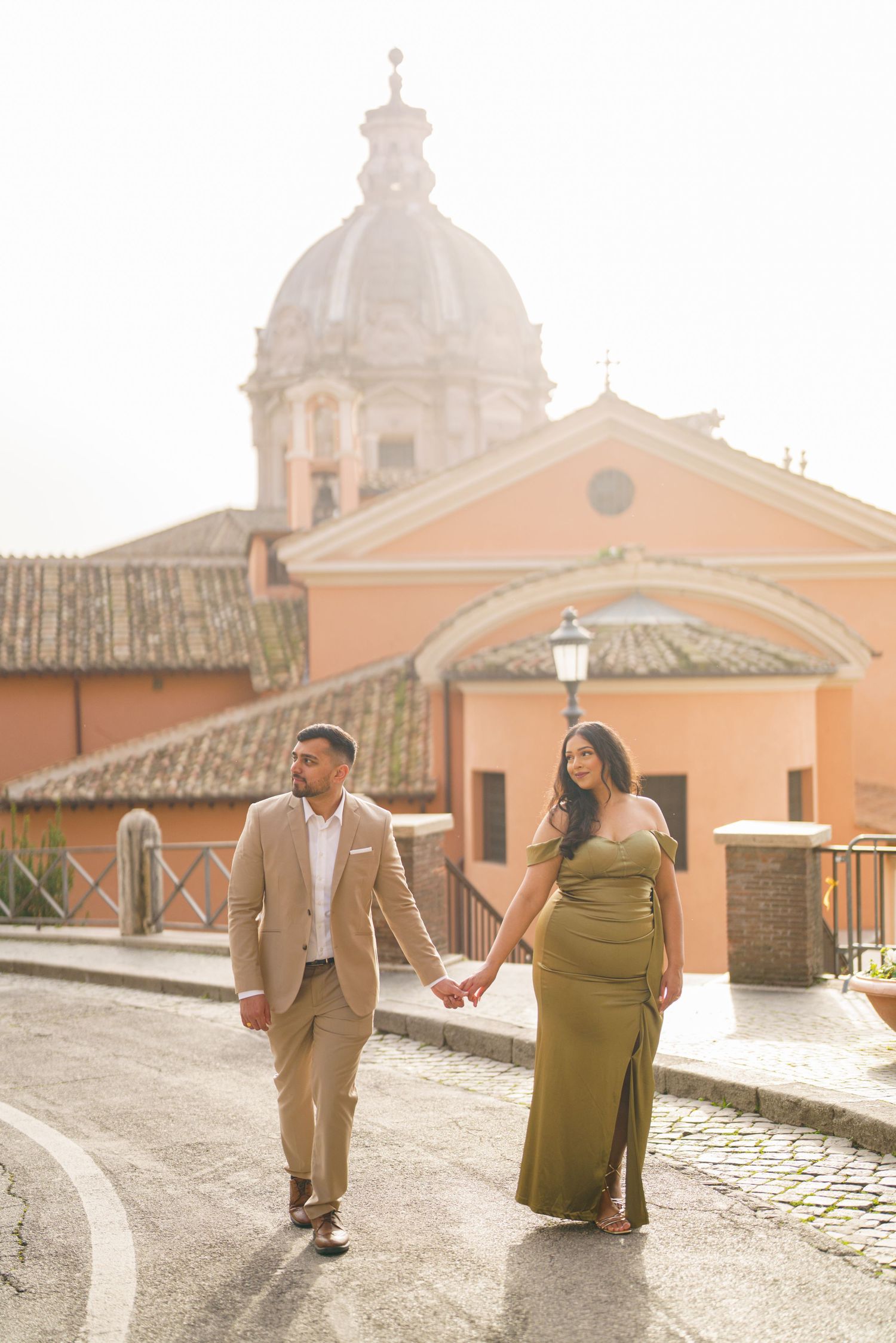 Couple holding hands walking on cobblestone street in Rome with historic church dome and warm golden light backdrop.