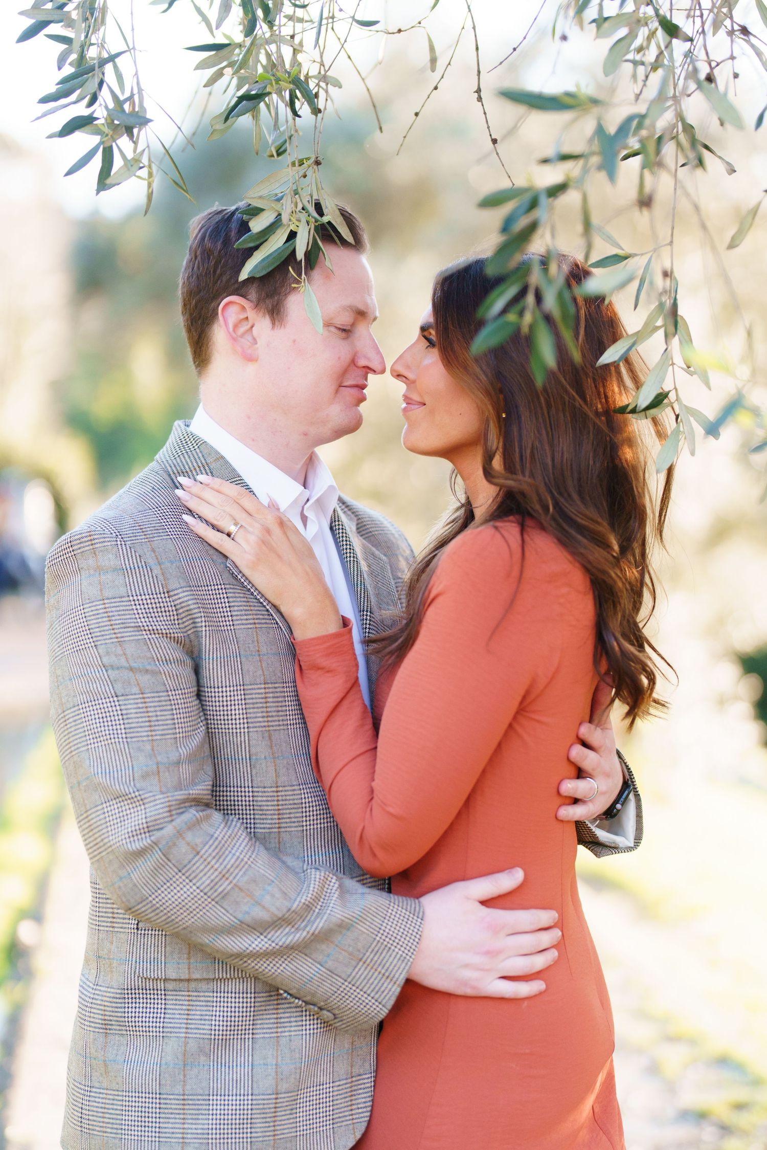 Couple sharing a romantic moment under olive branches, man in plaid blazer and woman in coral dress embracing outdoors.