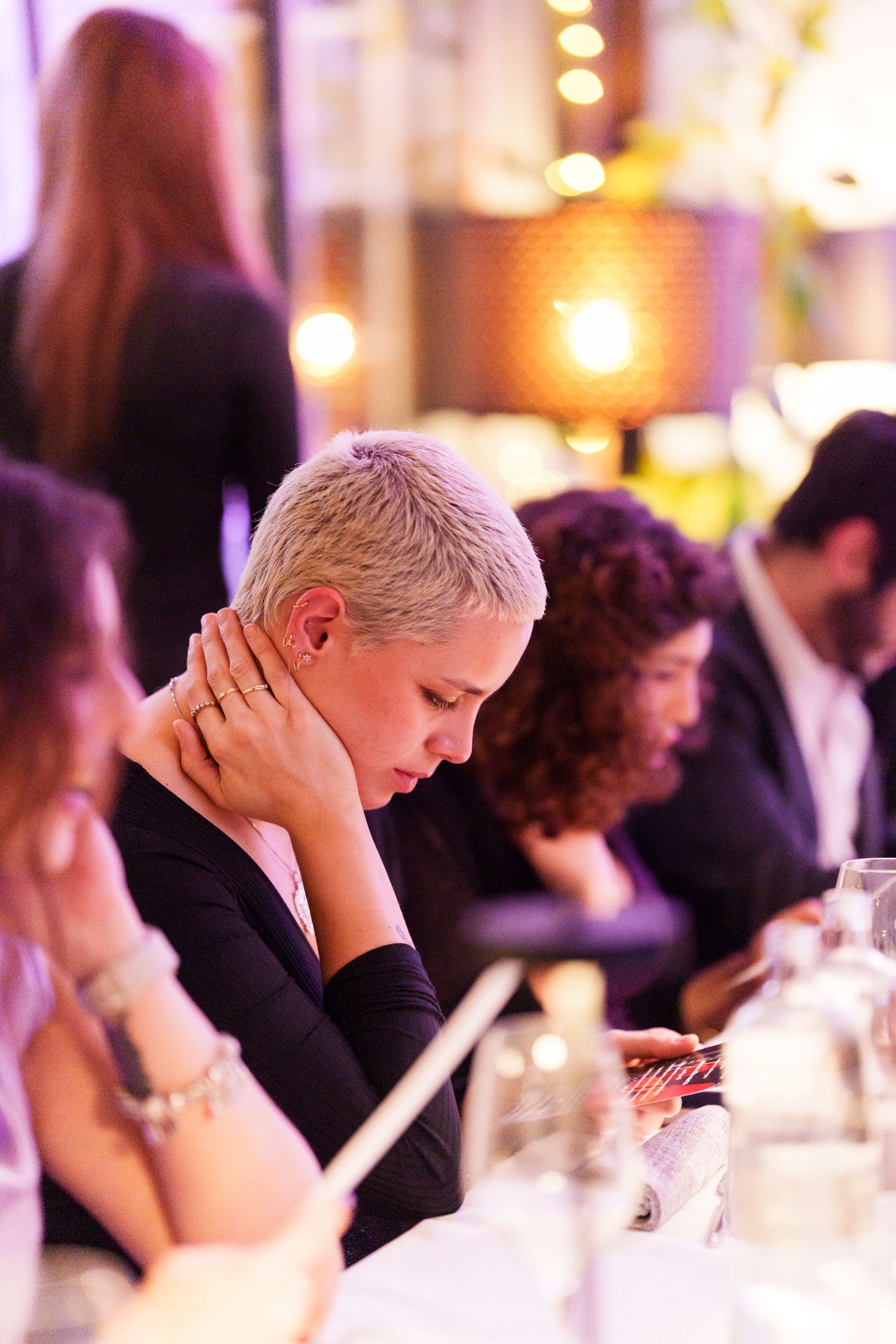 Woman with short blonde hair in black top looks down at phone during elegant dinner event with warm lighting.