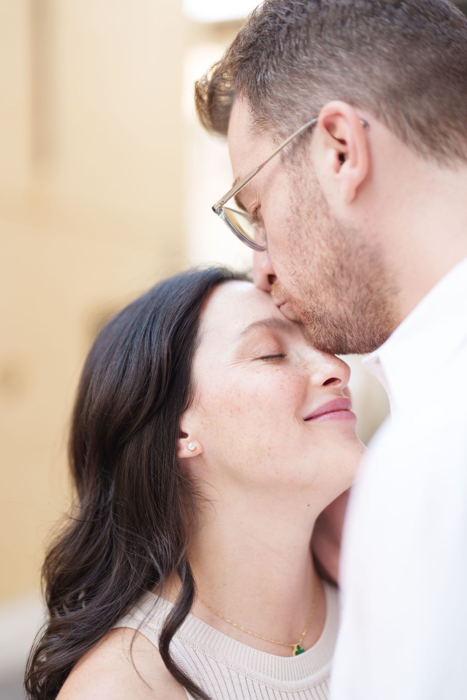 A man with glasses kissing a smiling dark-haired woman on the forehead in a tender romantic moment.