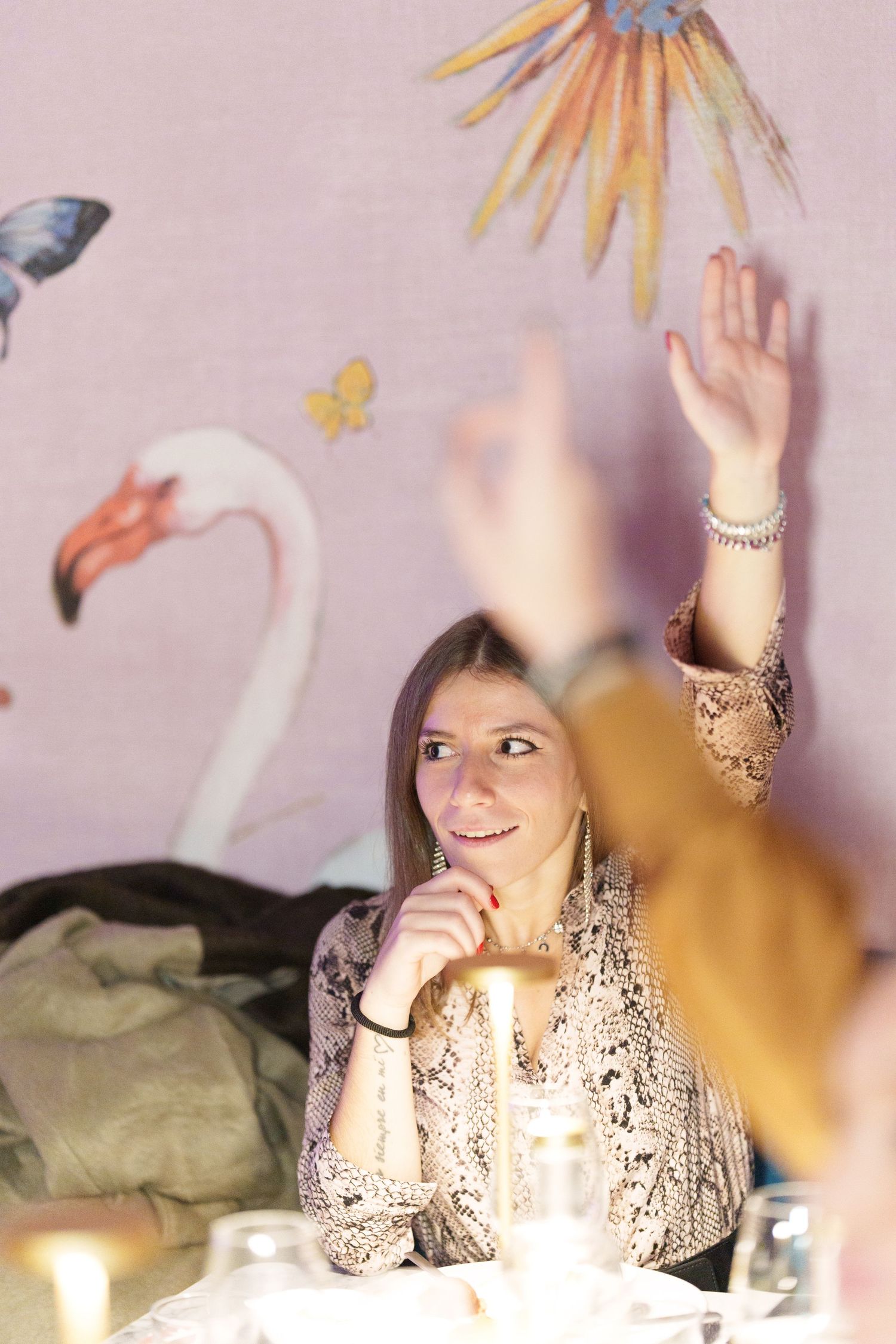Woman smiling at a dinner table with whimsical flamingo wallpaper while another person raises their hand nearby.
