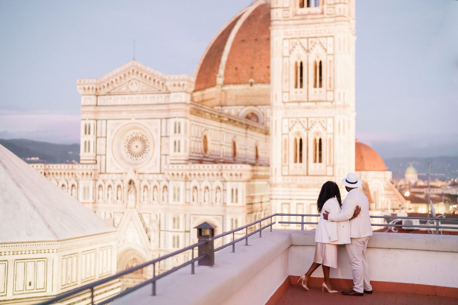 Couple in white attire embracing on a rooftop terrace overlooking Florence Cathedral dome at sunset.