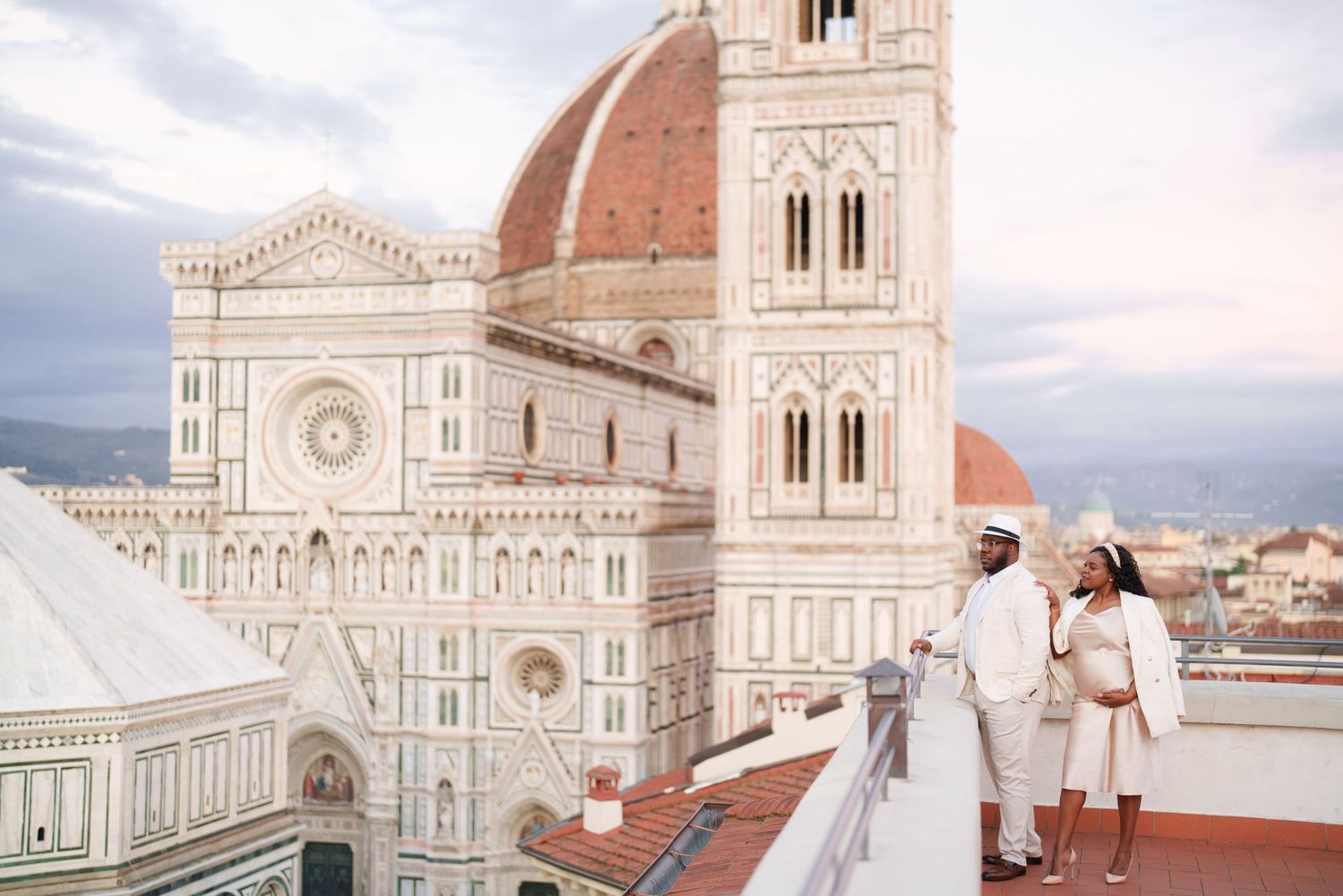 Couple posing on a rooftop terrace with Florence Cathedral dome and bell tower backdrop at dusk.