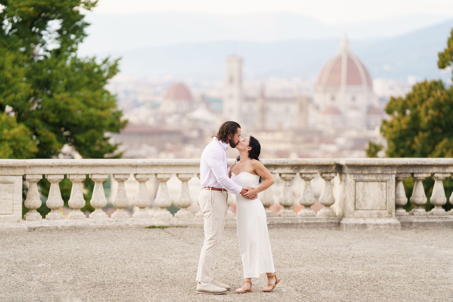 A couple shares a romantic kiss at a scenic overlook with Florence's iconic Duomo cathedral visible in the background.