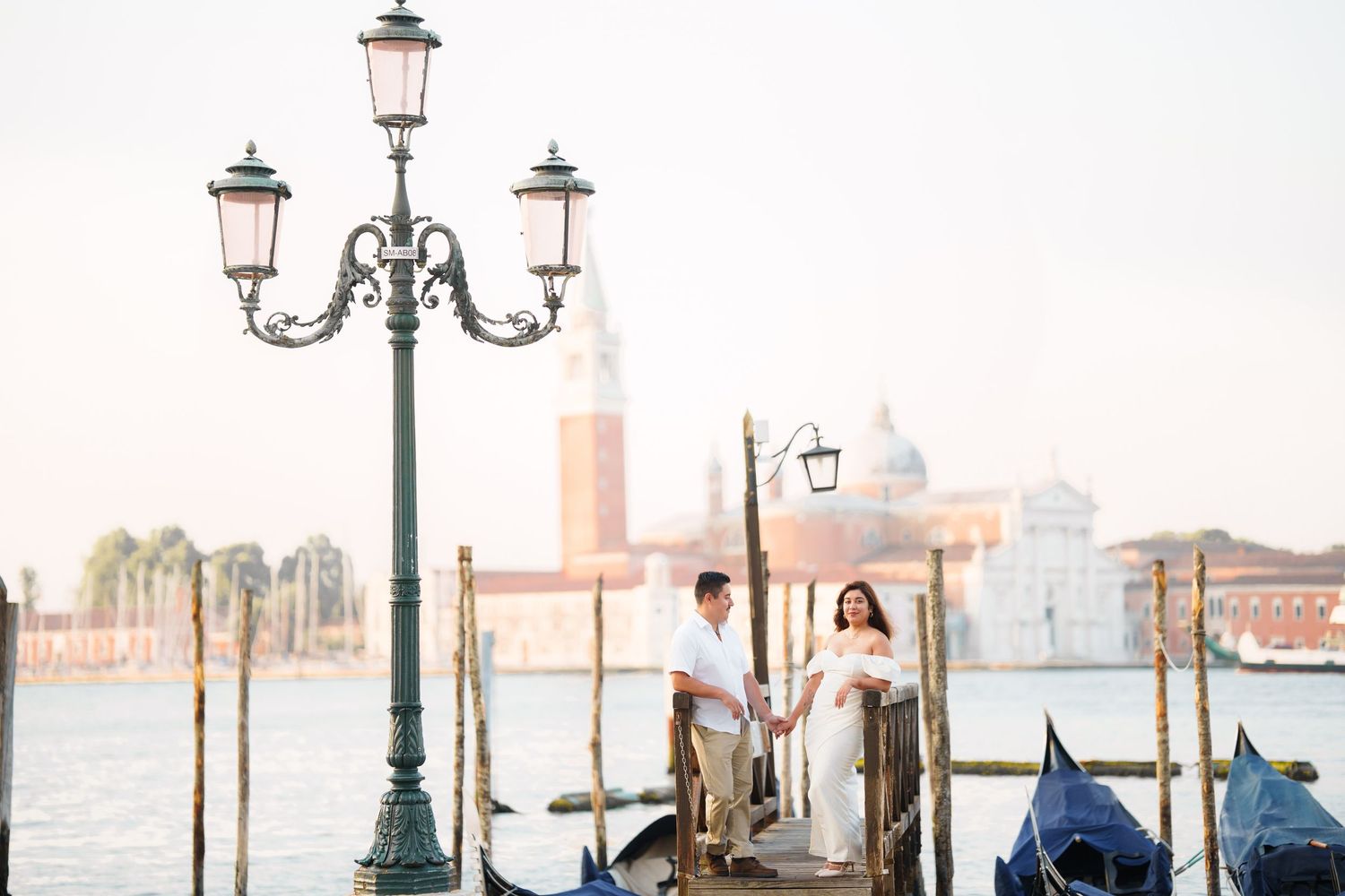 Ornate vintage street lamps line a misty waterfront in Venice with historic buildings visible in the background.