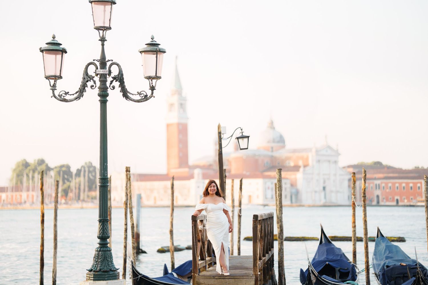 Venetian waterfront with ornate lampposts, gondolas, and the San Giorgio Maggiore church in the misty background.